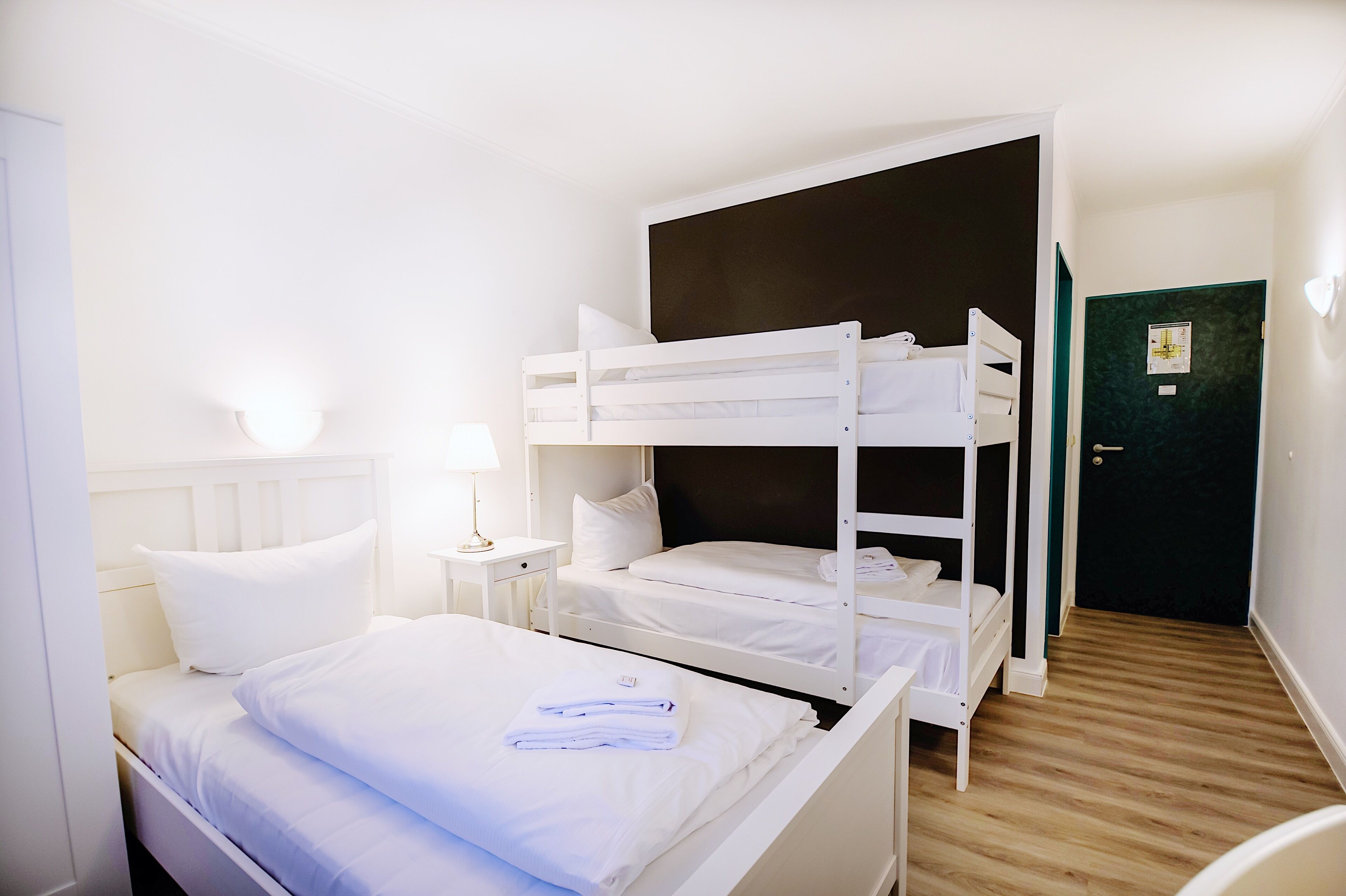 A room with a bunk bed, a bedside table with a lamp, and a door with a green frame.