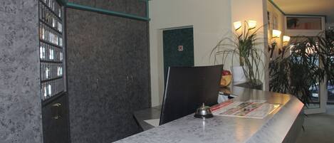 Una reception con un bancone in marmo, un monitor nero e un codice QR sul piano di lavoro. A destra, una pianta e sullo sfondo una porta con un cartello di divieto di accesso.