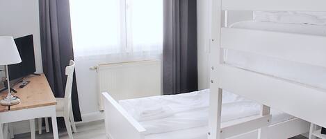 Una cama litera blanca con escritorio y silla, un radiador y una ventana con cortinas.