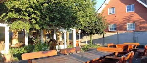 A courtyard with wooden picnic tables and benches, surrounded by a brick building and a large tree.