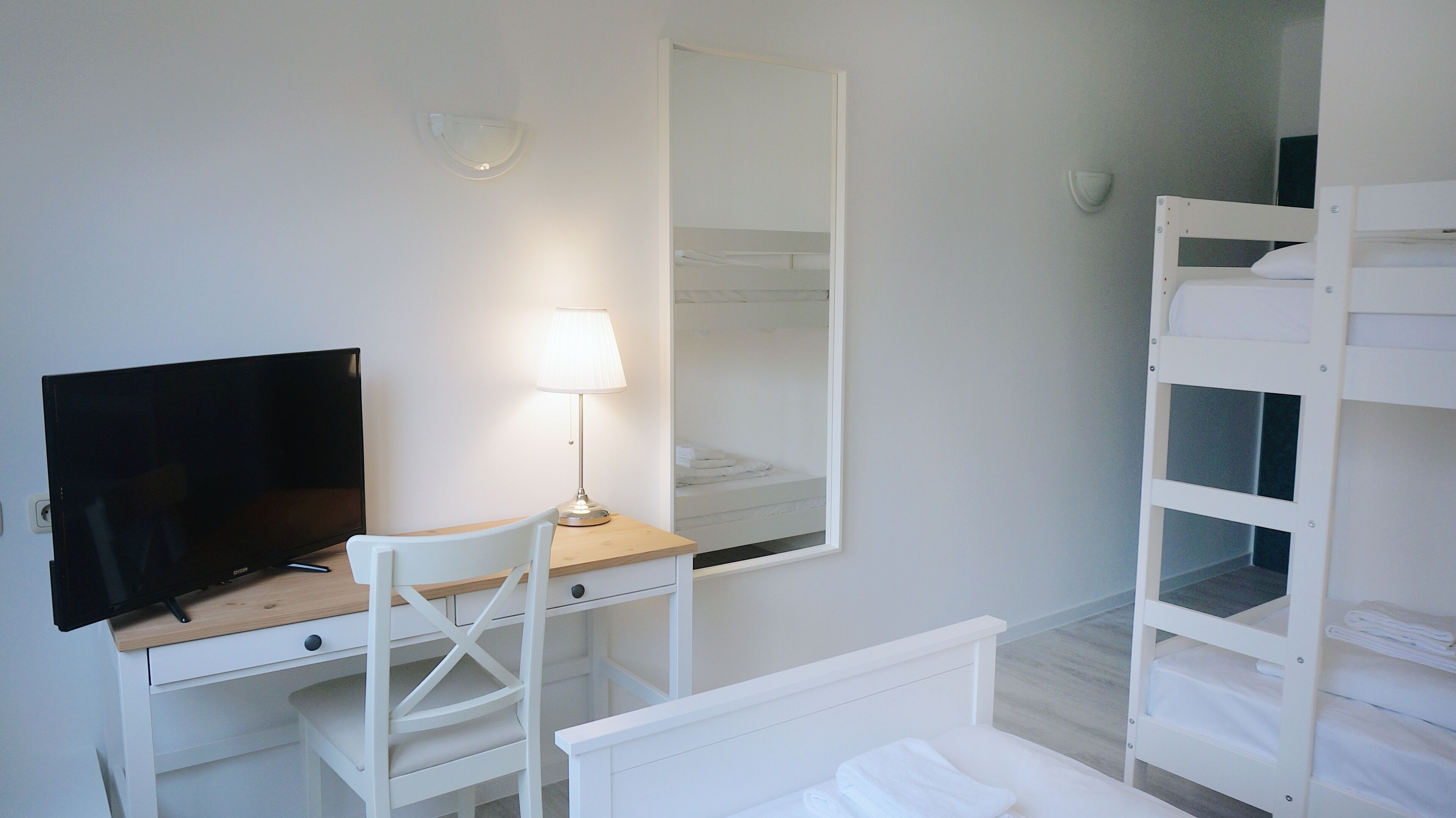 A bedroom with a single bed, a desk with a TV, a chair, a mirror, and a bunk bed.