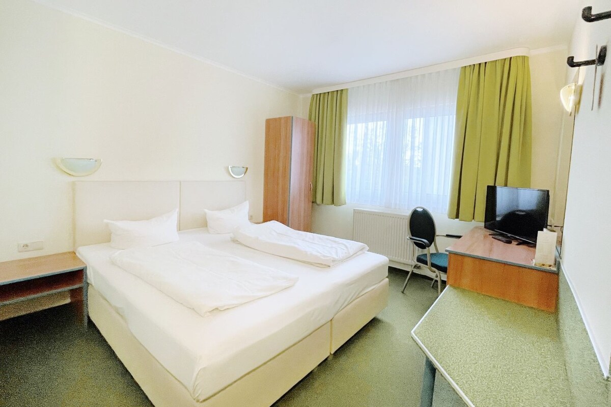 Habitación de hotel con una cama individual, un escritorio con televisor, una silla y una ventana con cortinas.