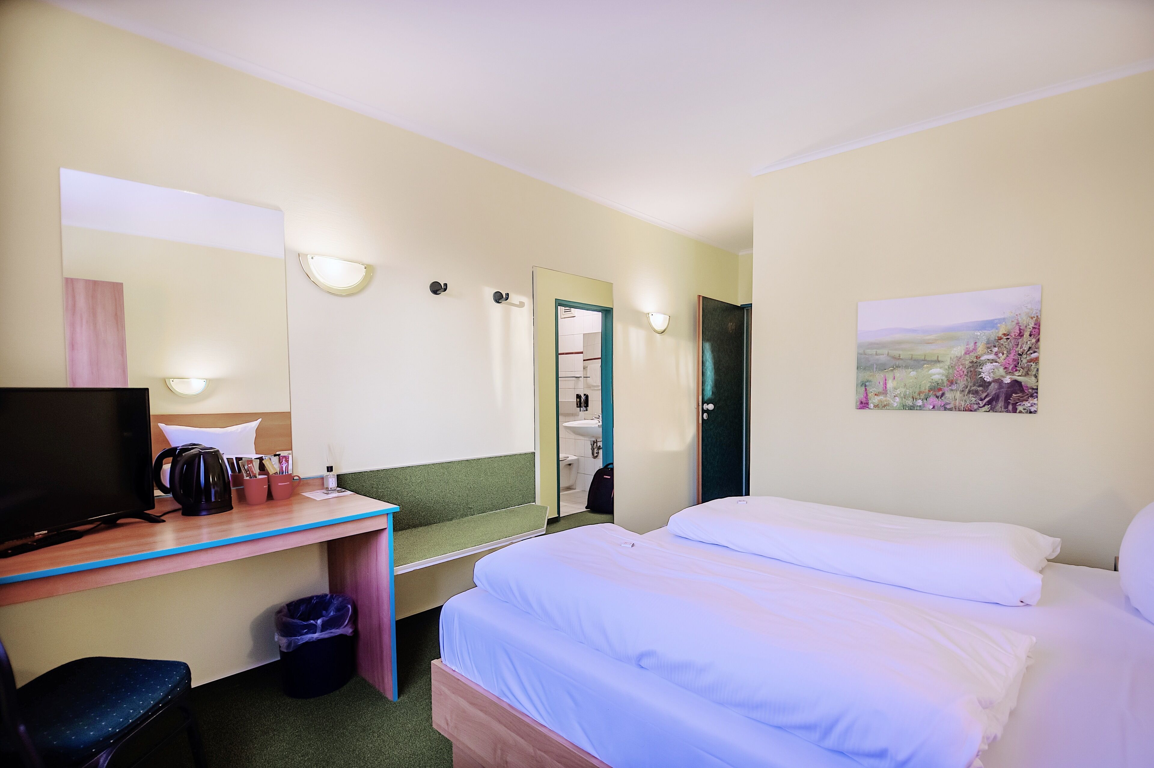 A hotel room with a bed, a desk with a computer, a chair, a trash bin, a mirror, and a painting on the wall.