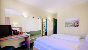 A hotel room with a bed, a desk with a computer, a chair, a trash bin, a mirror, and a painting on the wall.