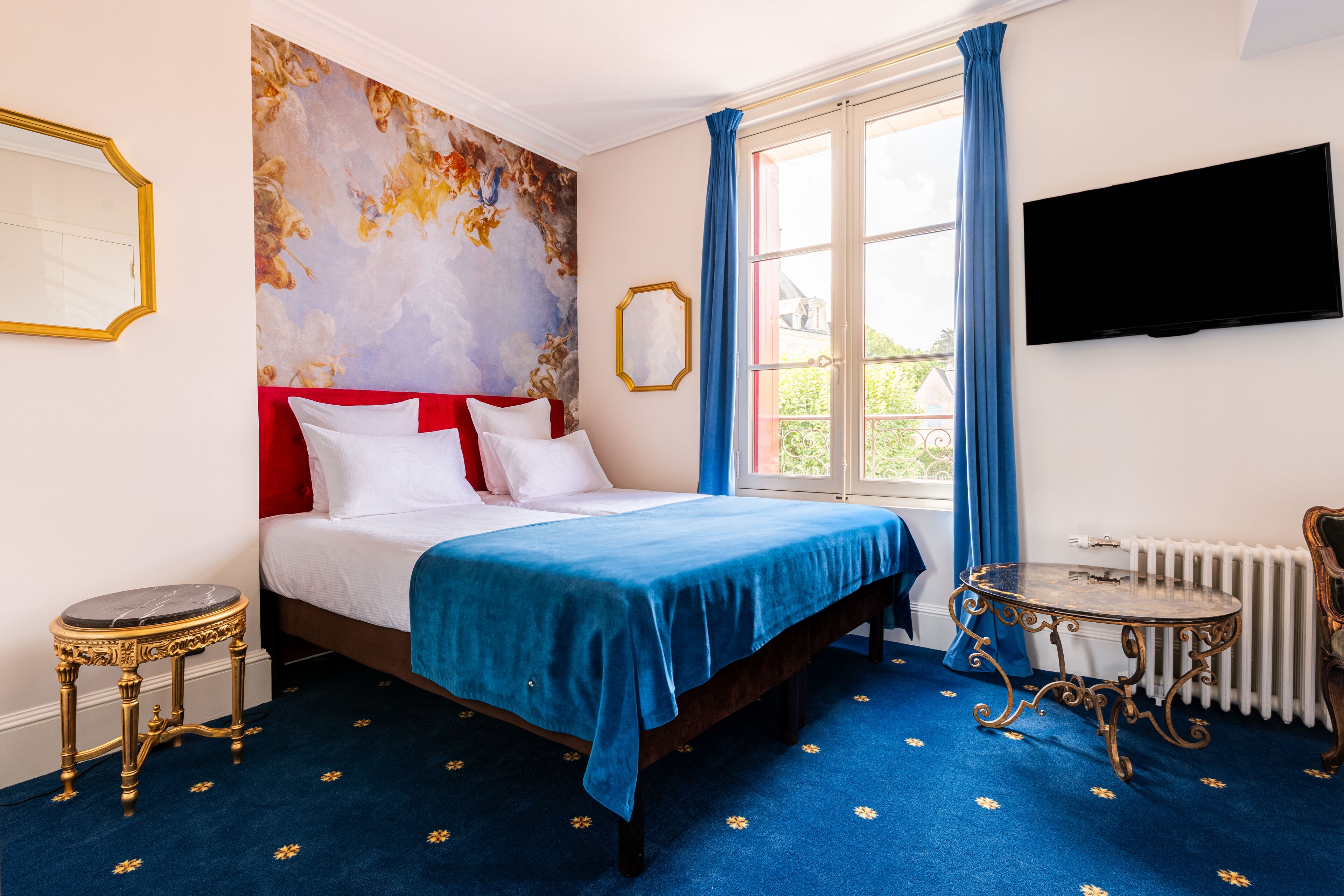 A bedroom with a bed, a blue carpet, a painting on the wall, a window with curtains, a radiator, and a television on the wall.