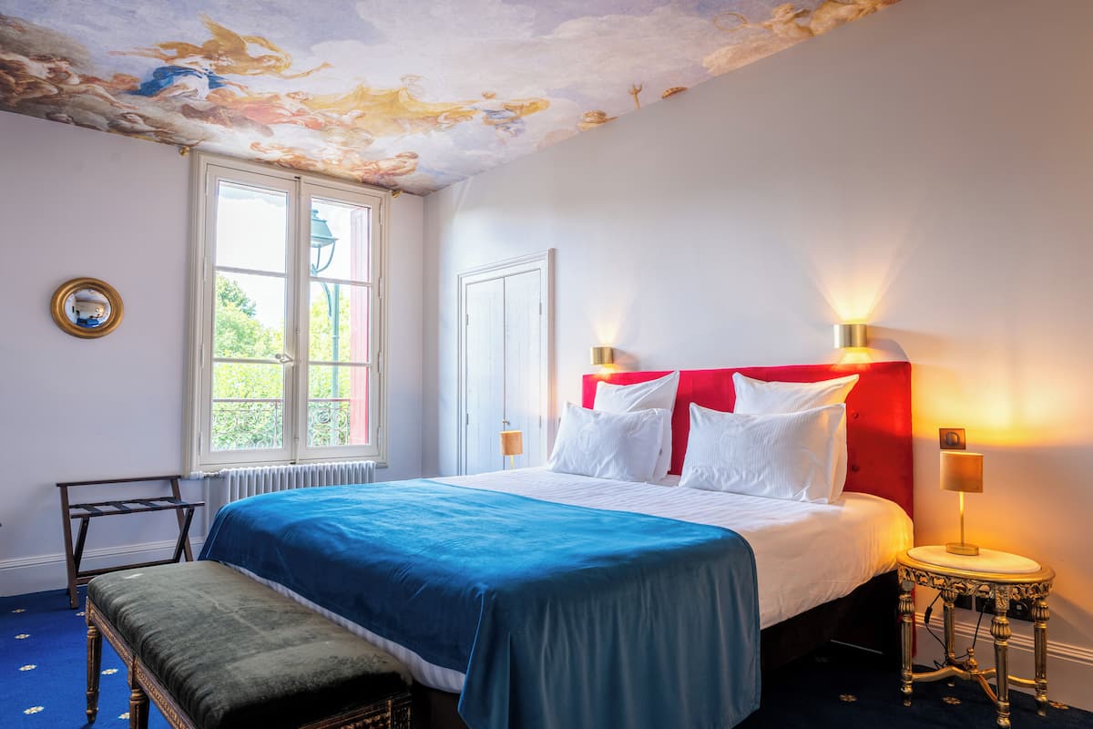 A hotel room with a bed, bench, lamp, and a window with a view of greenery.