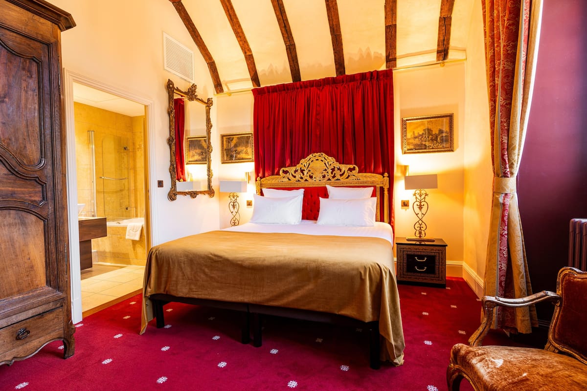 A hotel room with a large bed, a wooden wardrobe, a bathroom with a glass shower, and a red carpet.