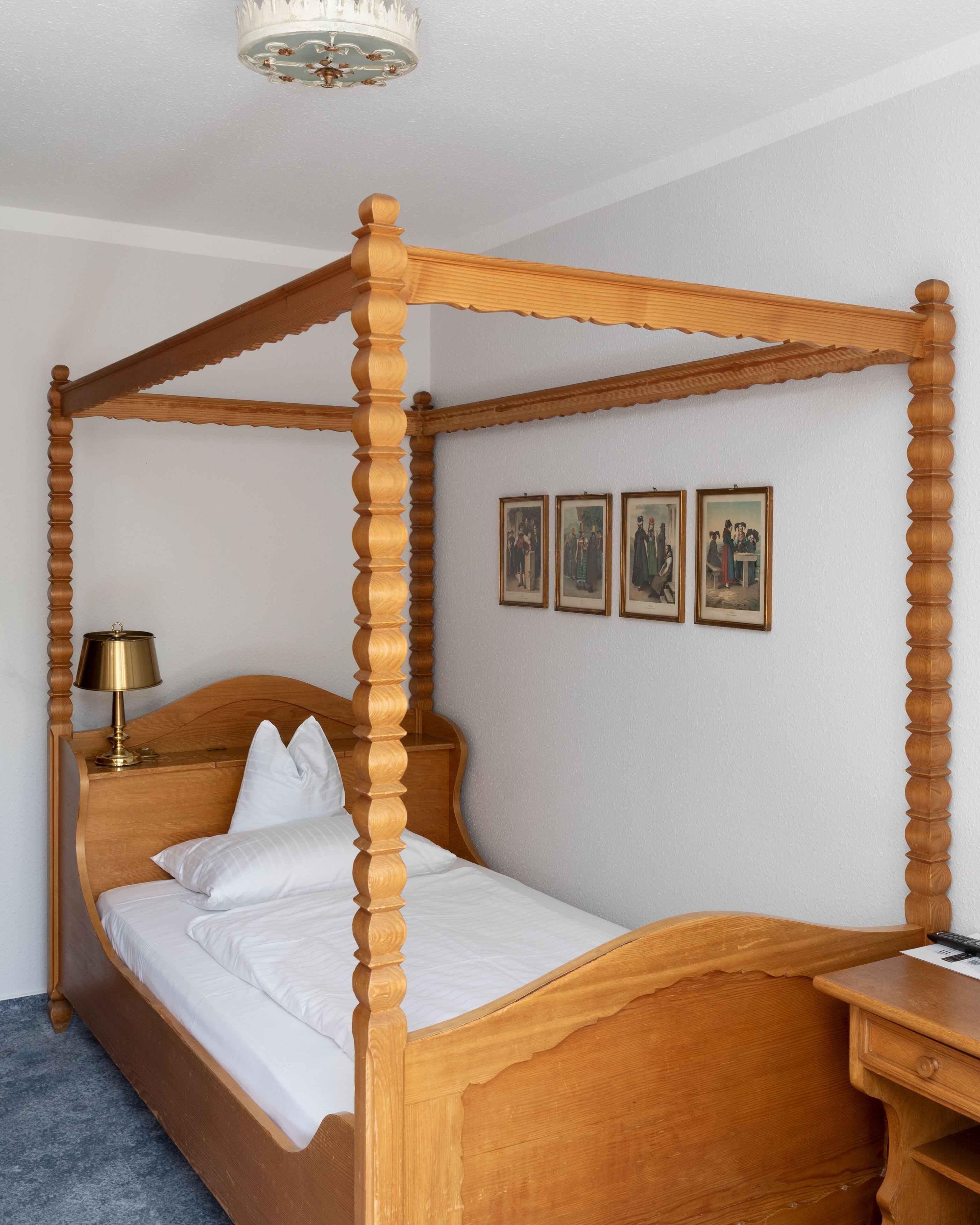 Una cama con dosel y largos postes, un cabecero de madera, una mesita de noche con lámpara y tres cuadros enmarcados en la pared.