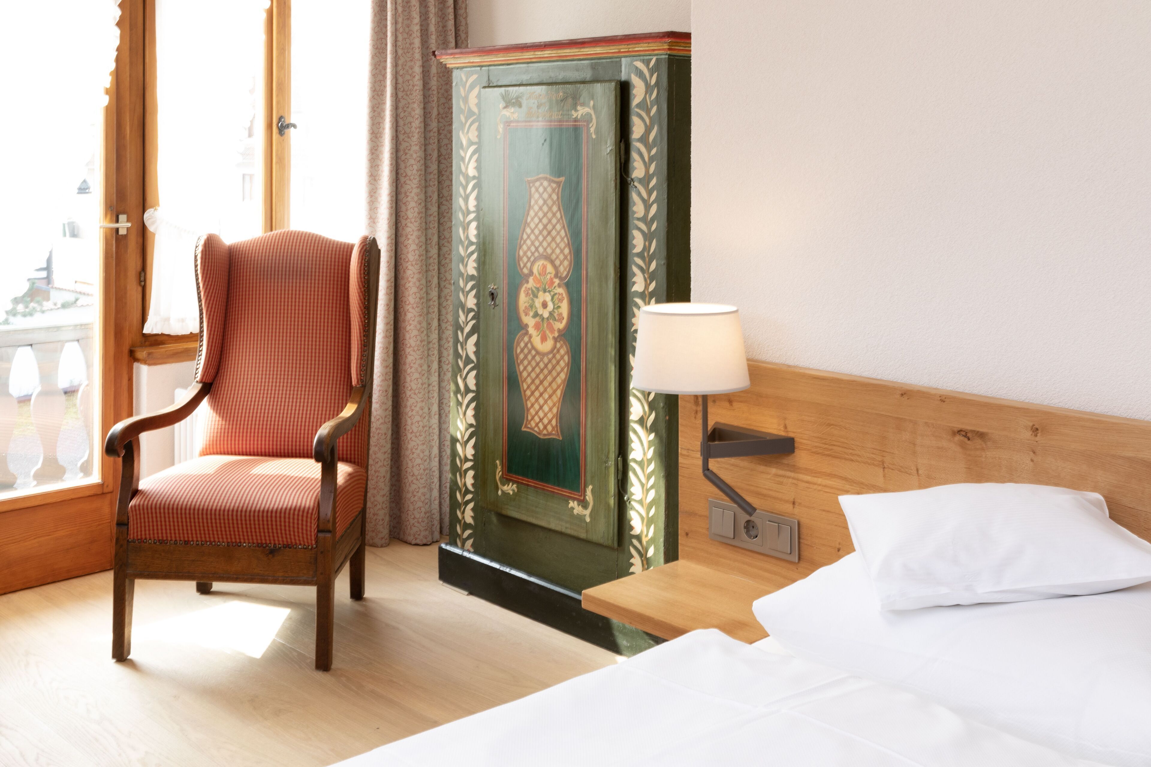 A hotel room with a wooden bed, a chair, a green cabinet, and a window with a view of a building.