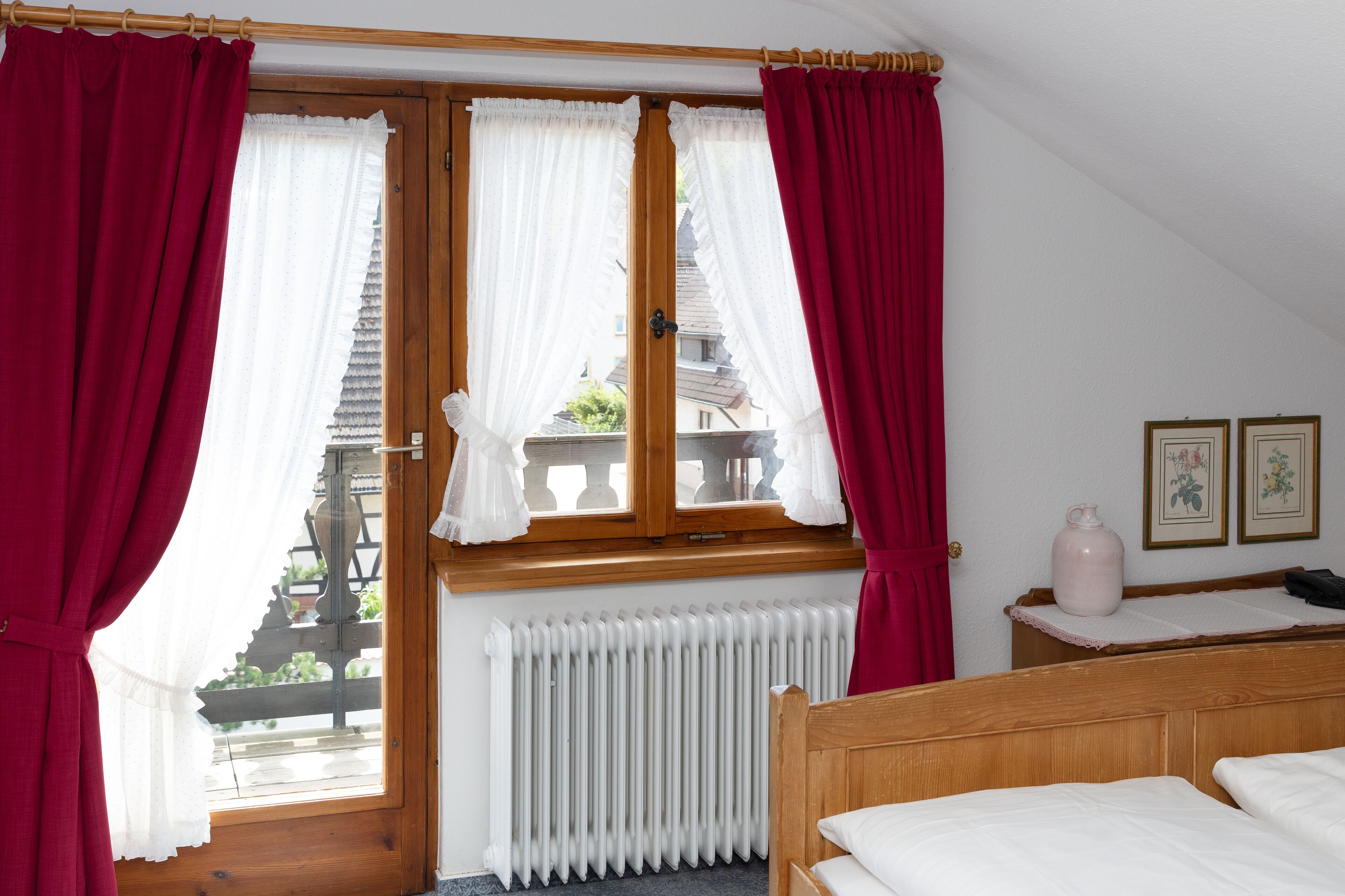 Ein Schlafzimmer mit einem hölzernen Bett, einem Heizkörper, einem Fenster mit Vorhängen und Blick auf einen Balkon.
