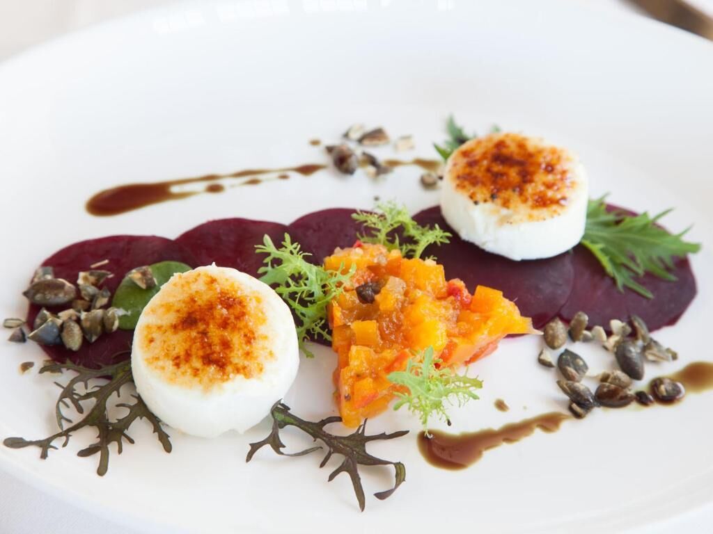A white plate with a dessert consisting of a white sphere, beetroot slices, and a mango mixture, garnished with seeds and greens.