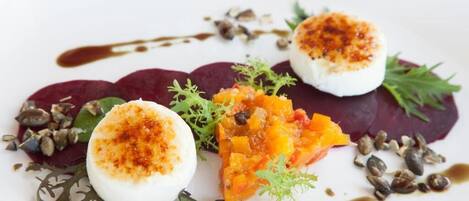 A white plate with a dessert consisting of a white sphere, beetroot slices, and a mango mixture, garnished with seeds and greens.