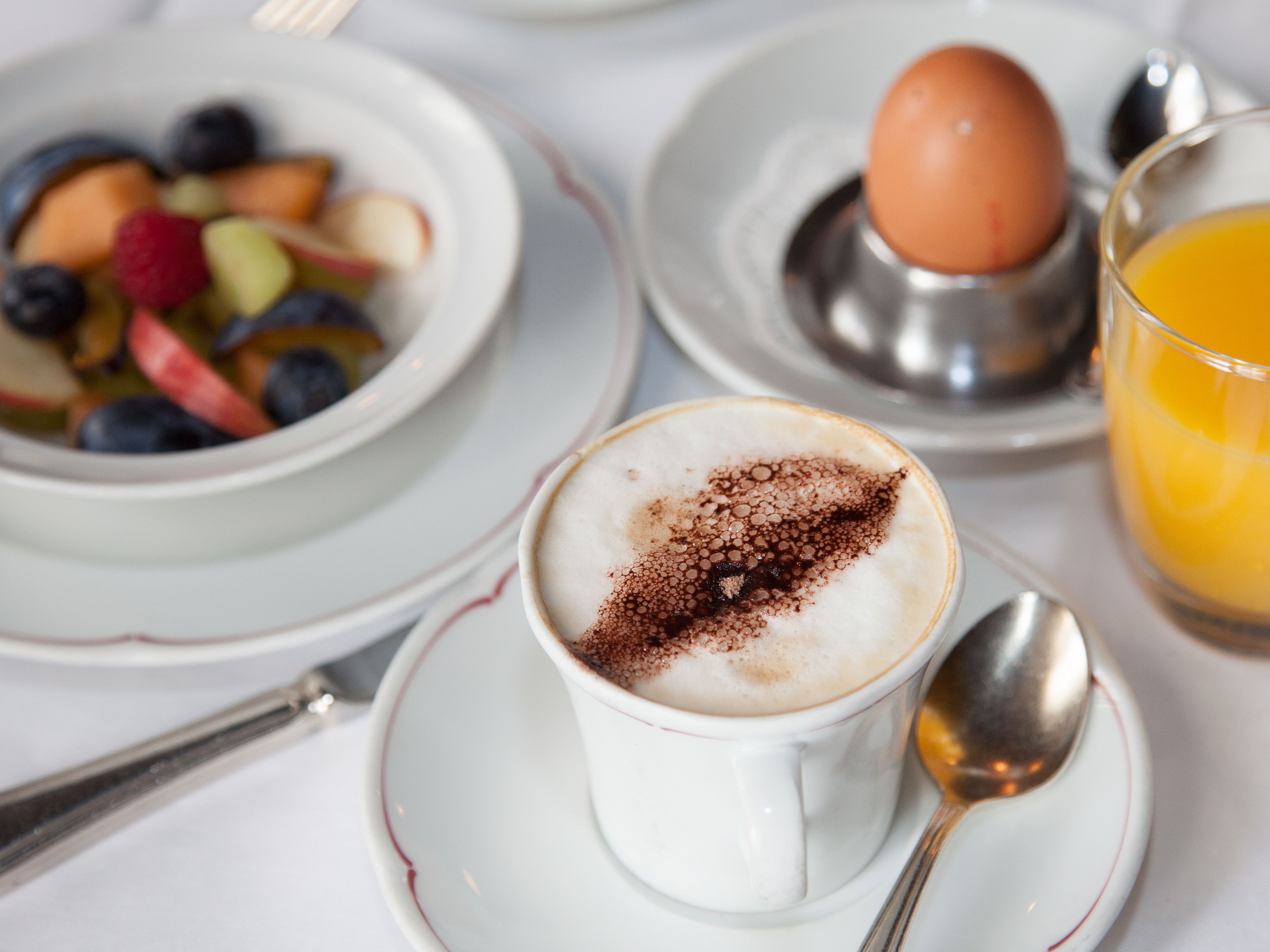 Ein Frühstückstisch mit einer Tasse Kaffee, Kakao, einer Fruchtschale, einem gekochten Ei und einem Glas Orangensaft.