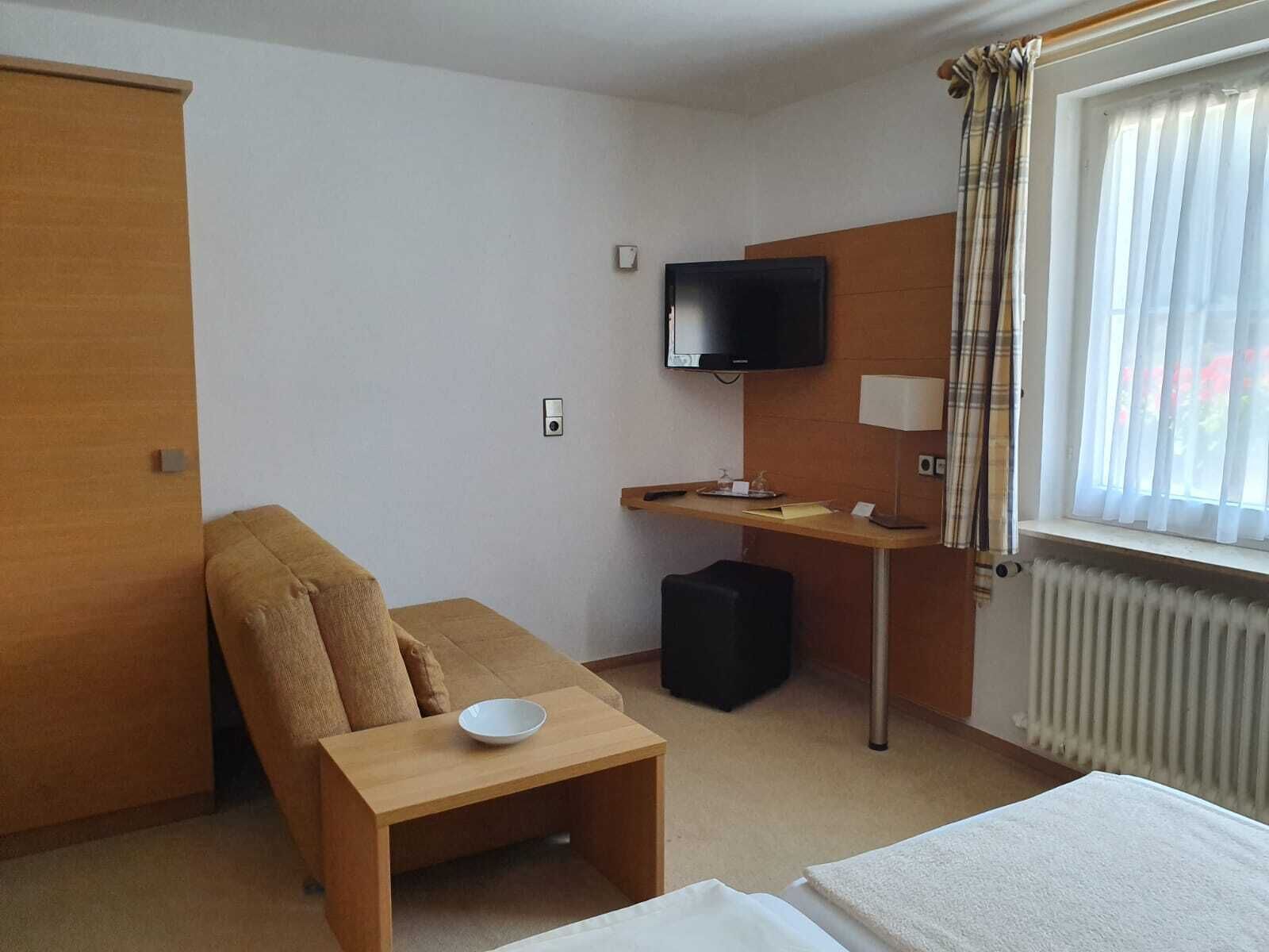 A hotel room with a bed, a small table with a bowl, a TV, a desk, a chair, a radiator, and a window with curtains.