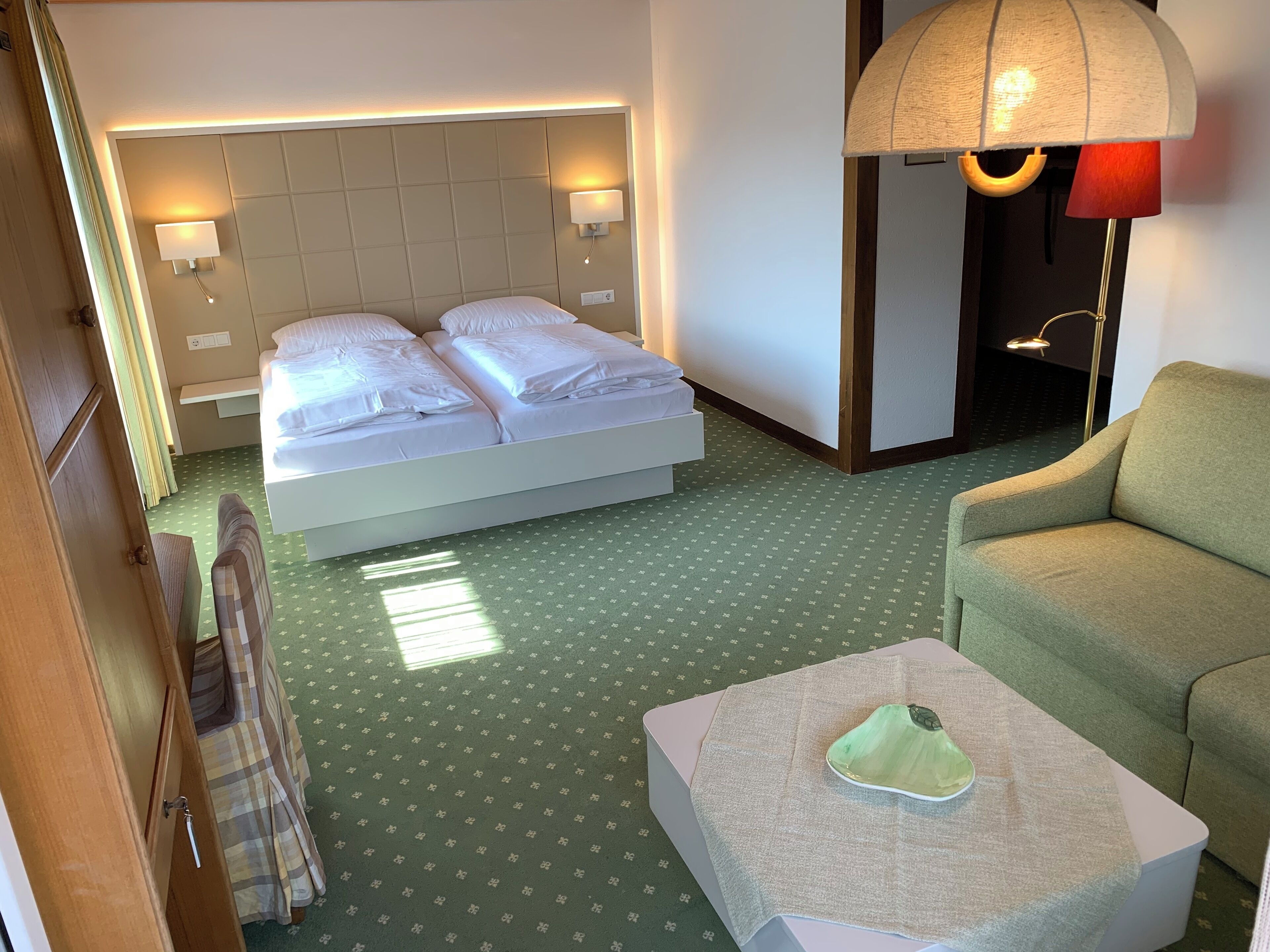 Una habitación de hotel con una cama, un sofá verde, una mesita con un objeto verde y una lámpara.