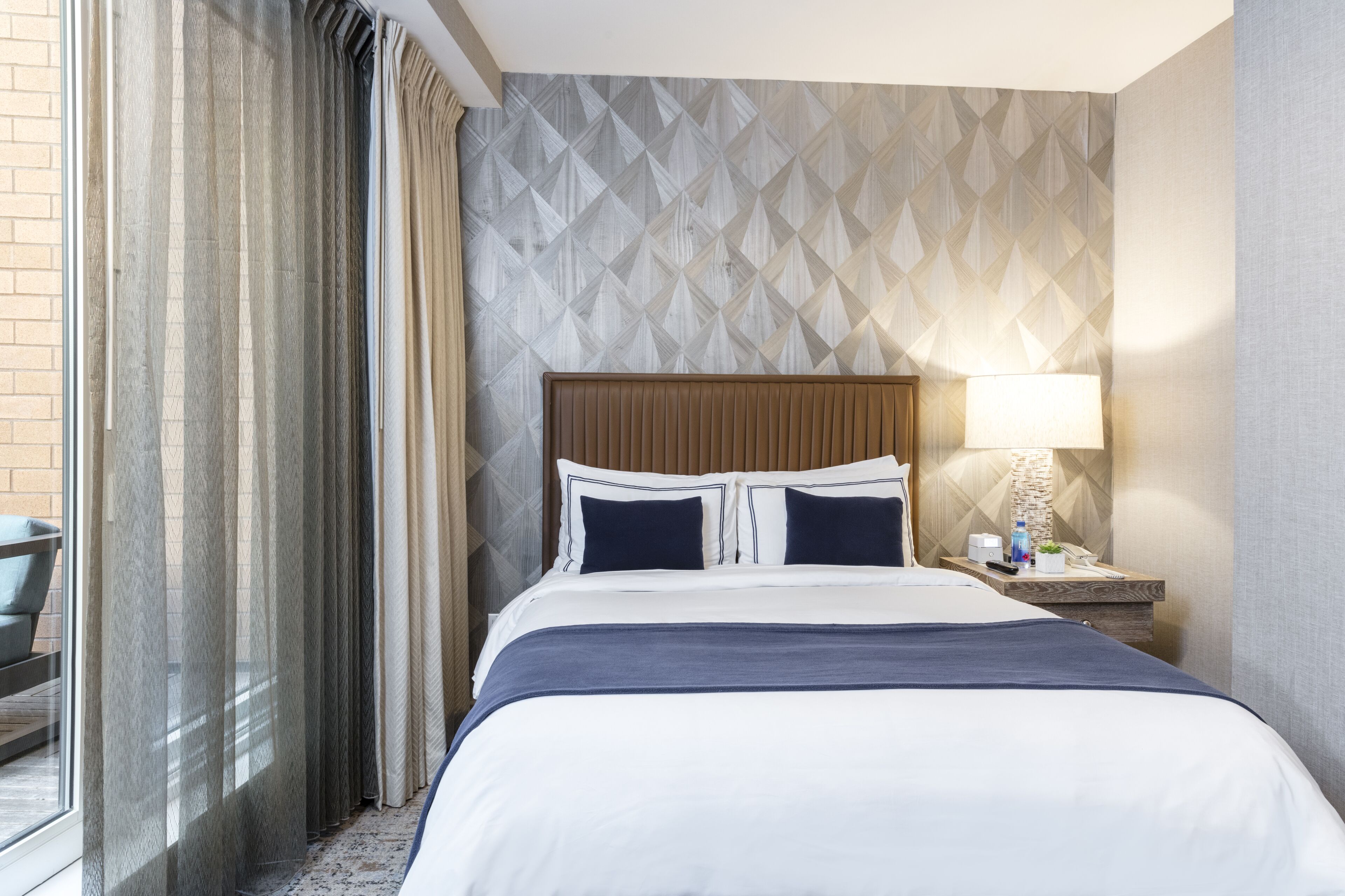 Een hotelkamer met een groot bed, een nachtkastje met een lamp, en uitzicht op de omgeving door een raam met gordijnen.