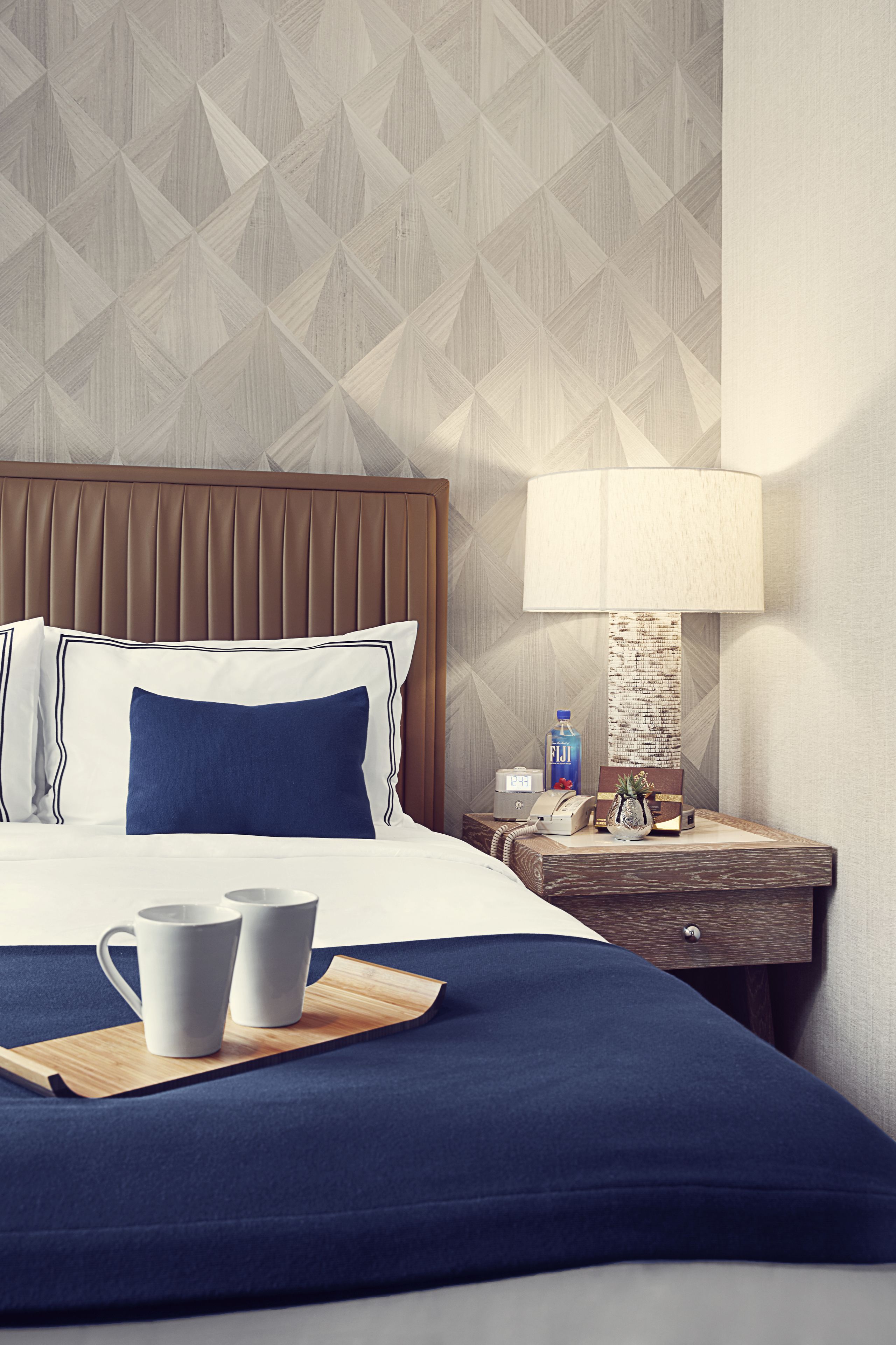 A hotel room with a bed, a nightstand, a lamp, and a tray with two mugs.