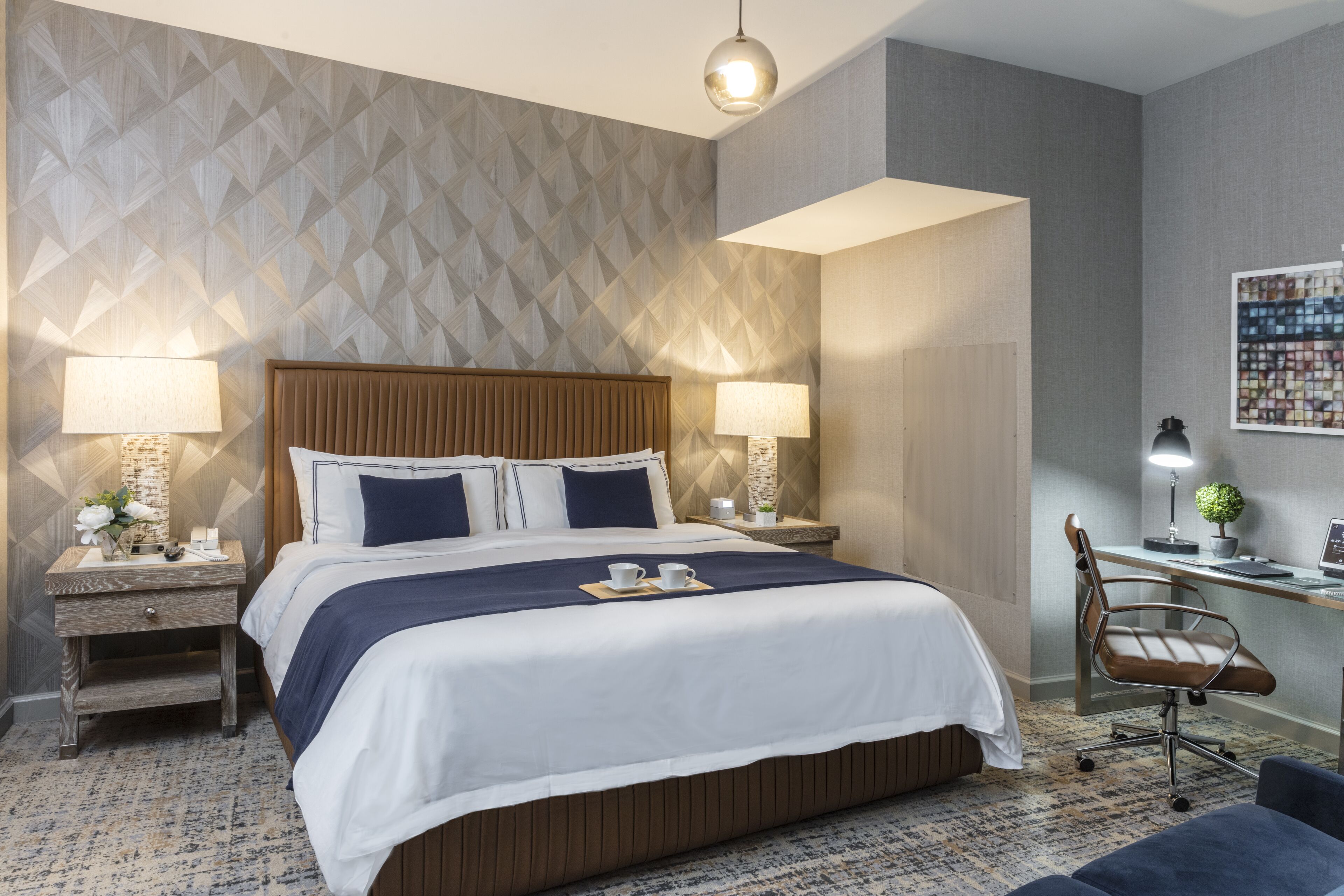 A hotel room with a large bed, two bedside tables with lamps, a desk with a chair, and a wall with a geometric pattern.