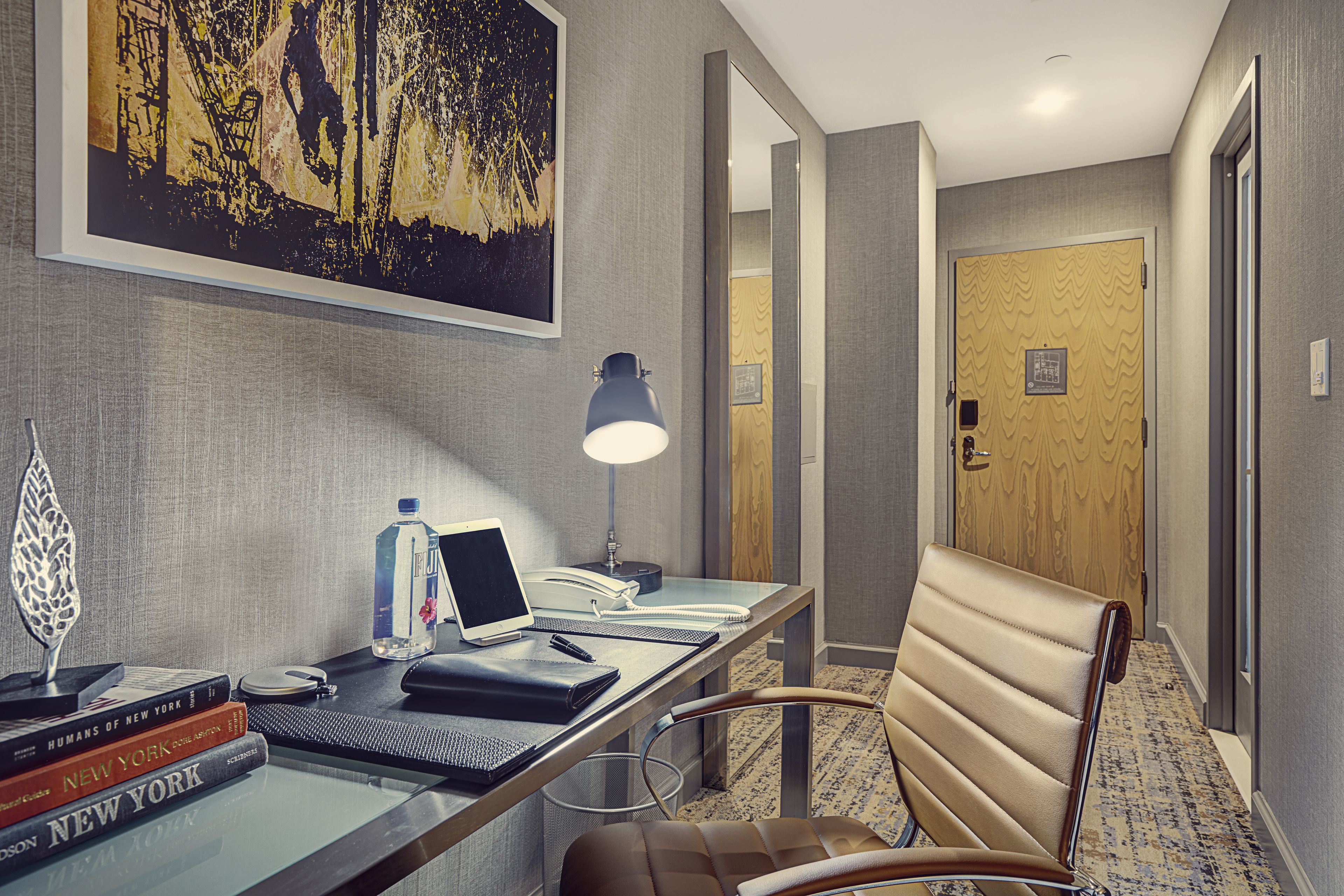 A hotel room with a desk, chair, lamp, books, and a painting on the wall.