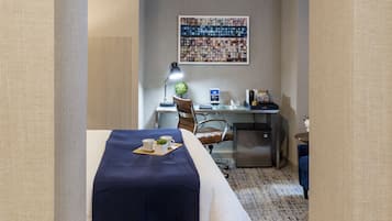 Une chambre d’hôtel comprenant un lit, un bureau et une chaise, une petite table avec une plante et une œuvre d’art accrochée au mur.