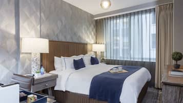 A hotel room with a large bed, a bedside table with a lamp, a small desk, and a window with curtains.