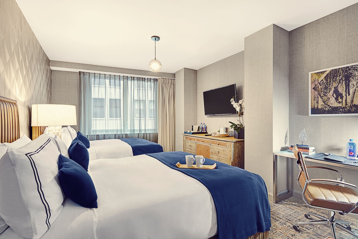 A hotel room with two beds, a desk, a chair, a TV, and a window with curtains.