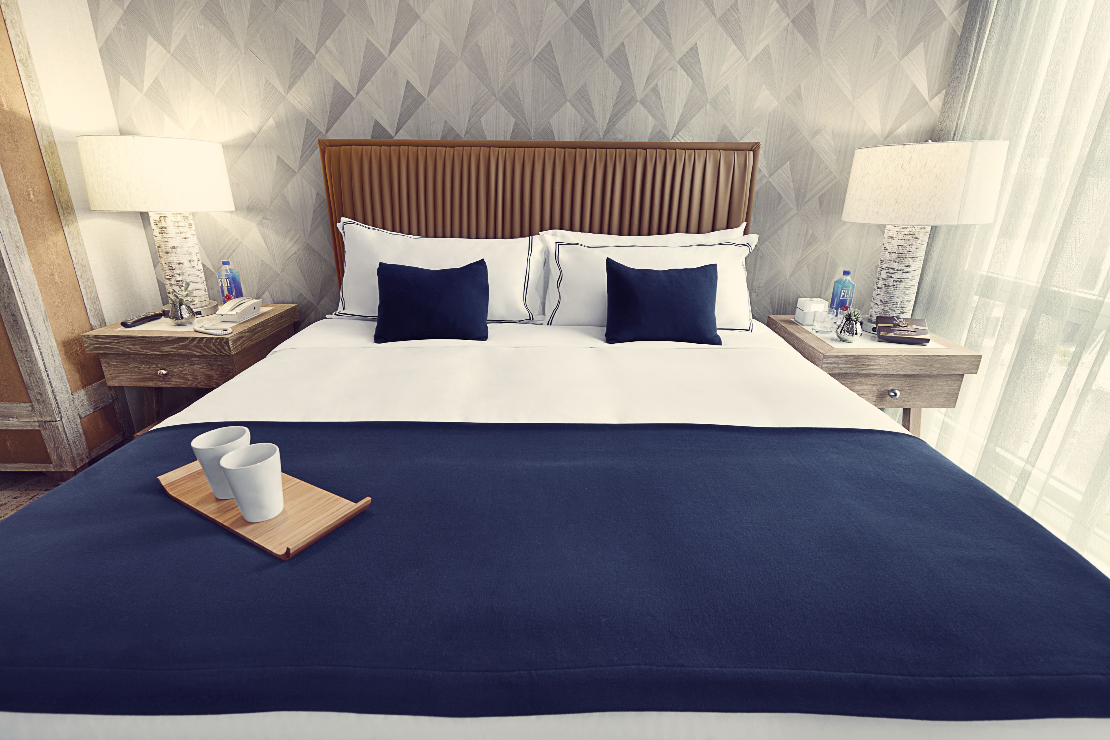 A hotel room with a large bed, two bedside tables, a wooden headboard, and a view of the city through sheer curtains.