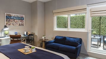 A hotel room with a bed, a blue sofa, a desk with a chair, a small round table with a plant, and a window with a view of greenery.