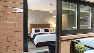 A hotel room with a bed, a small table, a chair, and a window with a view of a building.