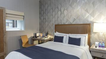 A hotel room with a large bed, a desk, a chair, and a nightstand with a lamp and water bottles.