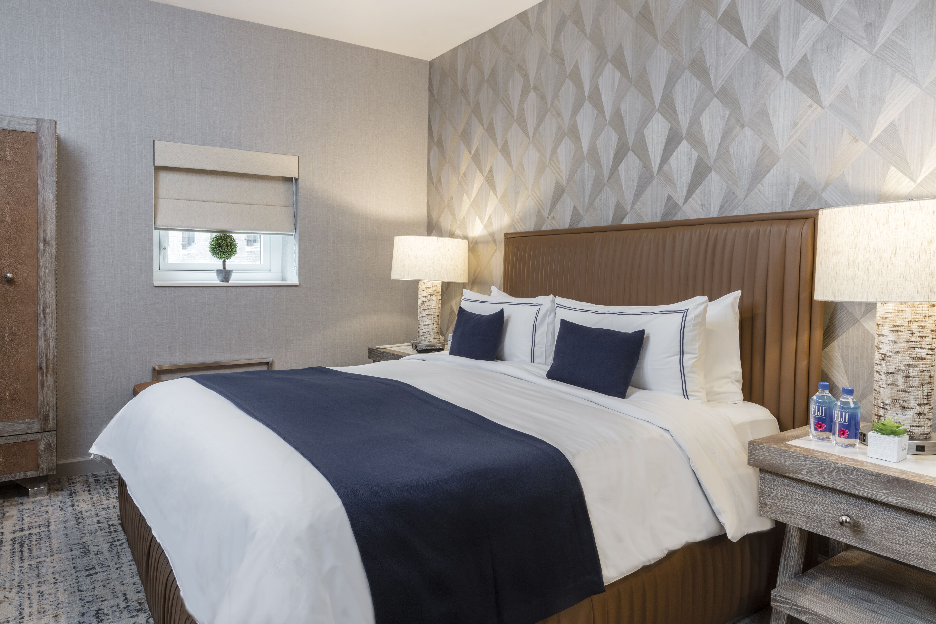 Quarto de hotel com uma cama grande, duas mesas de cabeceira, um guarda-roupa de madeira, uma janela com vista e uma parede com padrão.