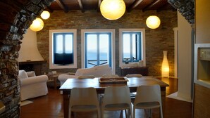A cozy dining area with a wooden table and white chairs, a stone fireplace, and large windows offering a view of the sea. - Etoile De Mer Villa (Andros)