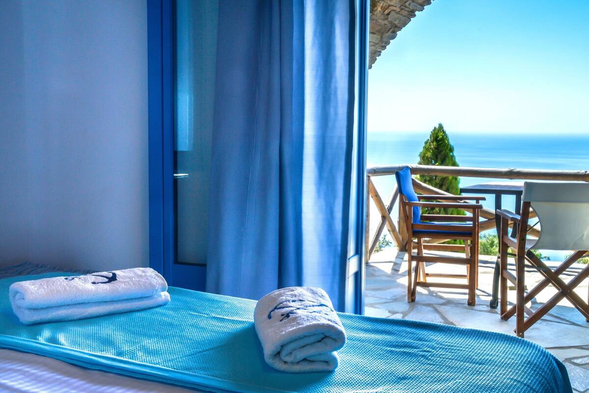 A room with a bed, a window with blue curtains, a view of the sea, and a table with chairs outside.