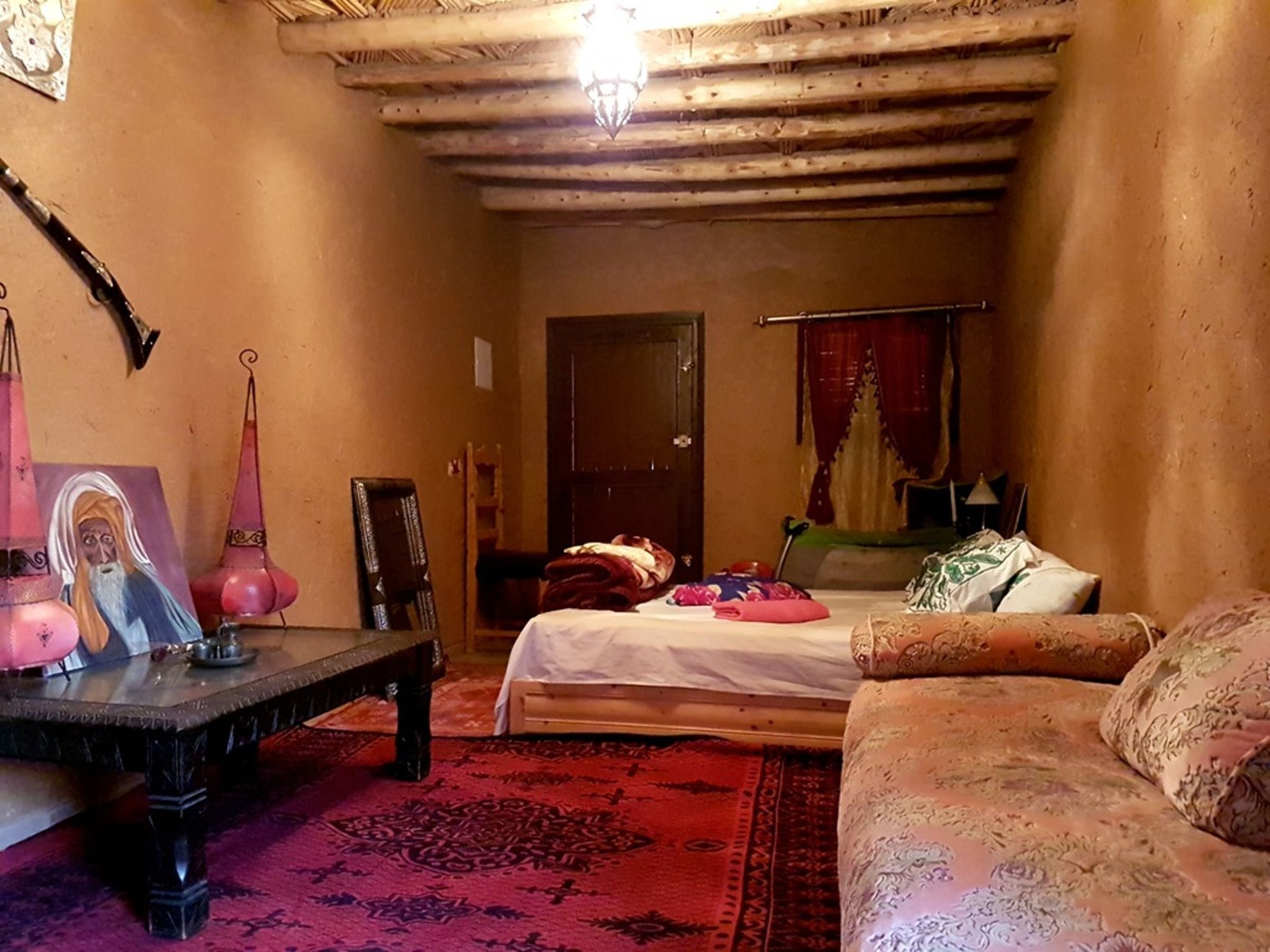 Quarto aconchegante com cama, sofá, mesa pequena e uma cadeira. Há uma porta ao fundo e um abajur pendurado.