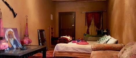 Quarto aconchegante com cama, sofá, mesa pequena e uma cadeira. Há uma porta ao fundo e um abajur pendurado.