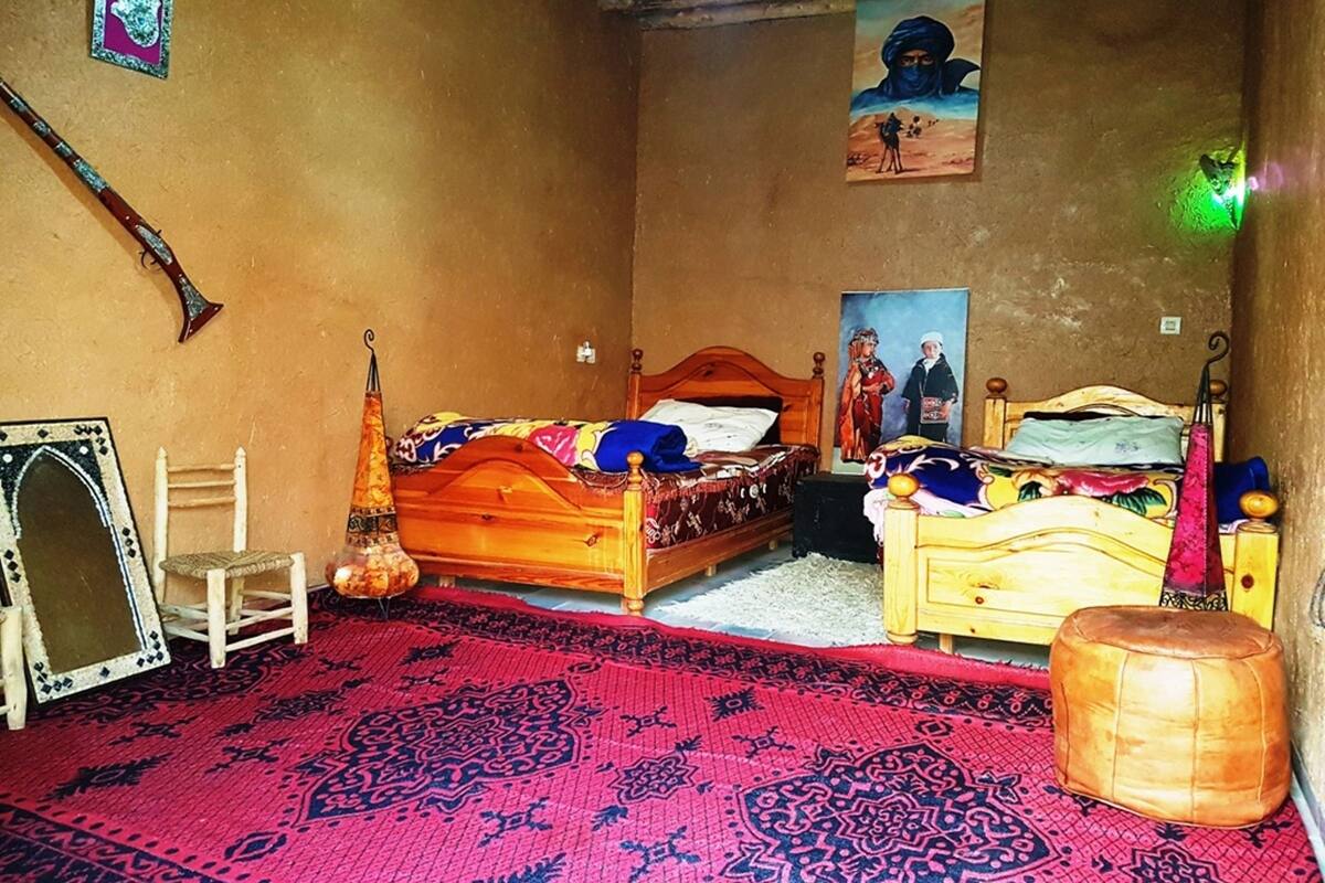 Una habitación con dos camas de madera, un tapete rojo con estampado, un taburete pequeño, una silla y una lámpara fijada a la pared.