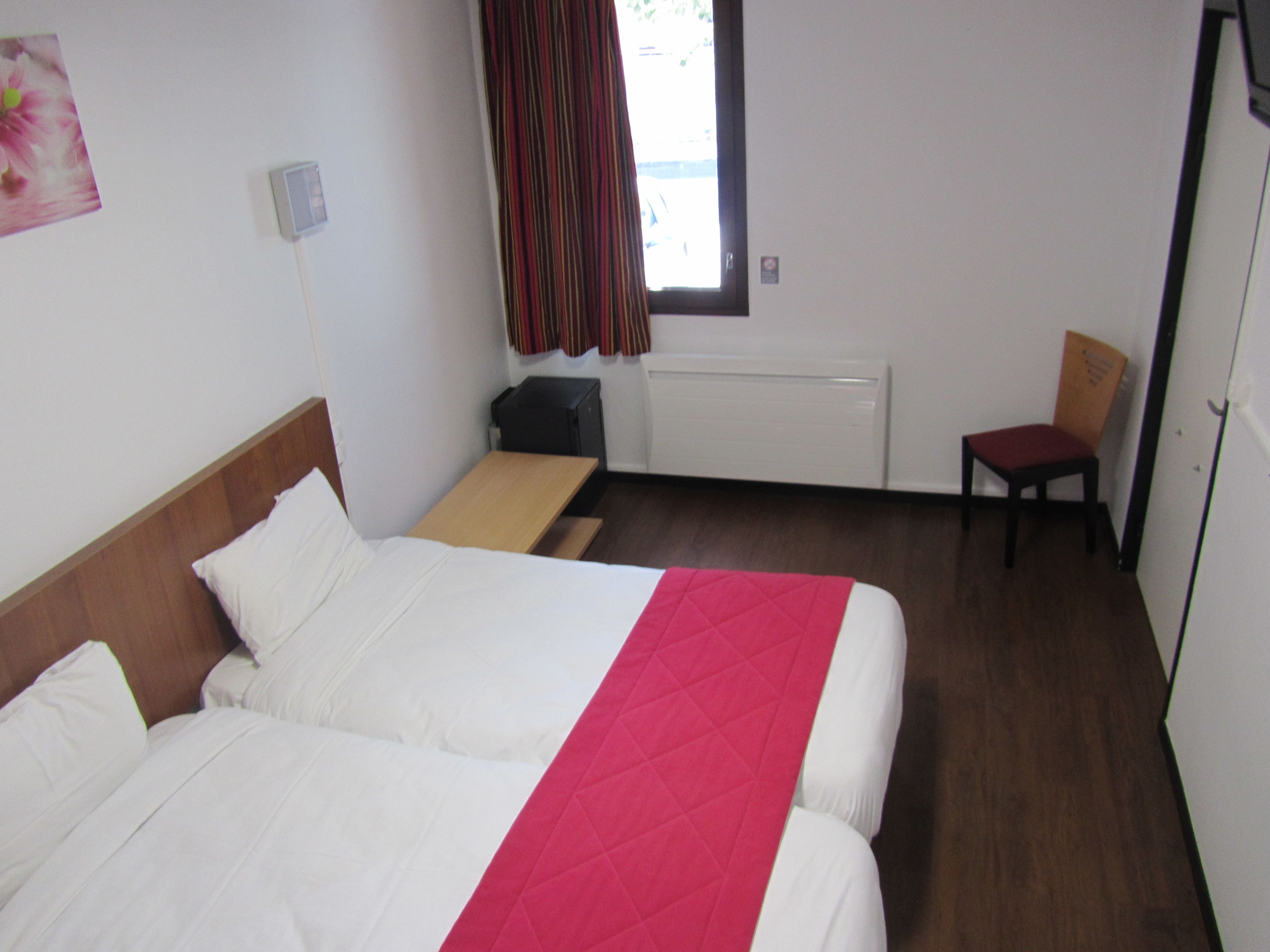 A hotel room with two beds, a desk, a chair, a radiator, and a window with curtains.