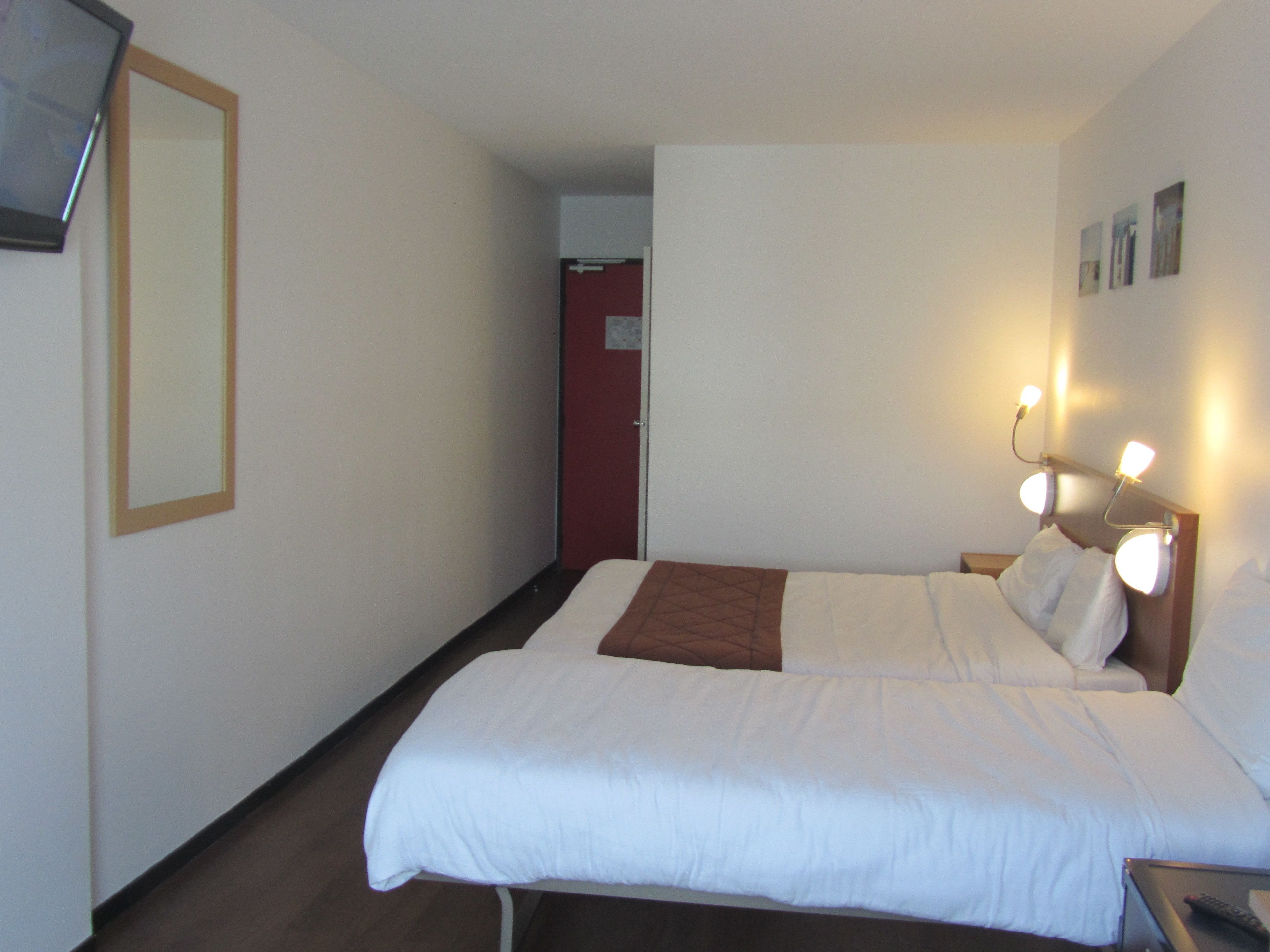 A hotel room with a bed, bedside table, wall-mounted lights, and a door leading to another room.