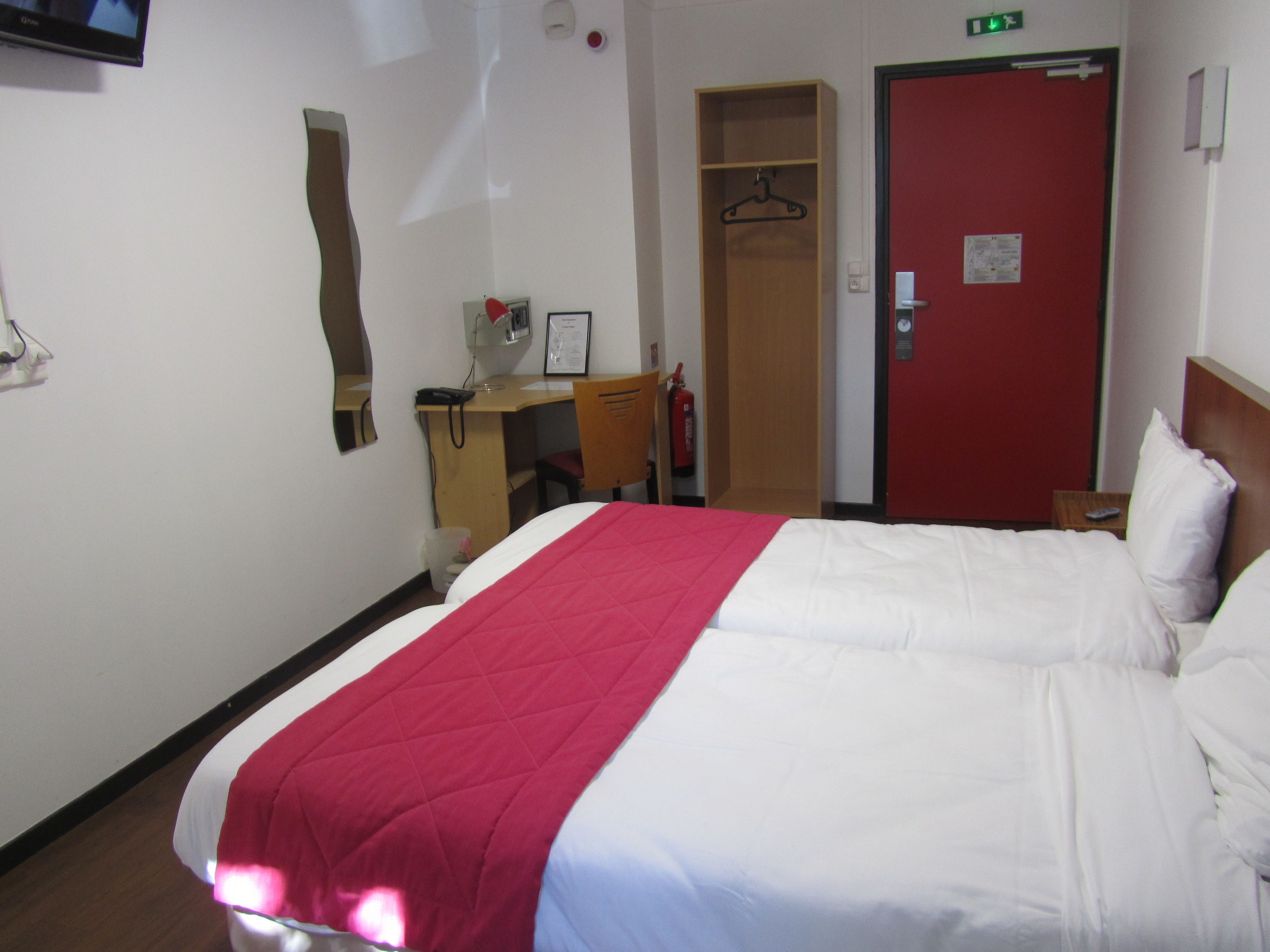 Habitación de hotel con cama, escritorio, silla, puerta roja y una mesita con control remoto.