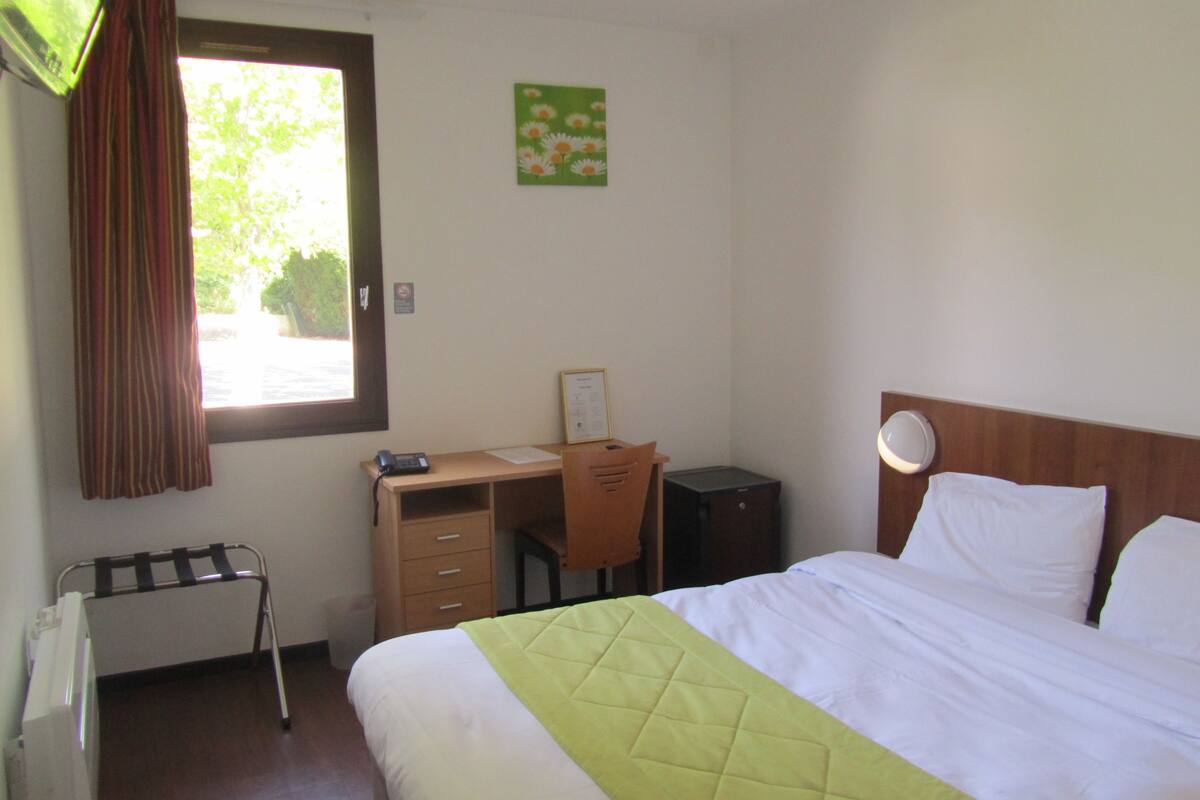Habitación de hotel con una cama, un escritorio con silla, una mesita, una lámpara de noche y una ventana con cortinas.
