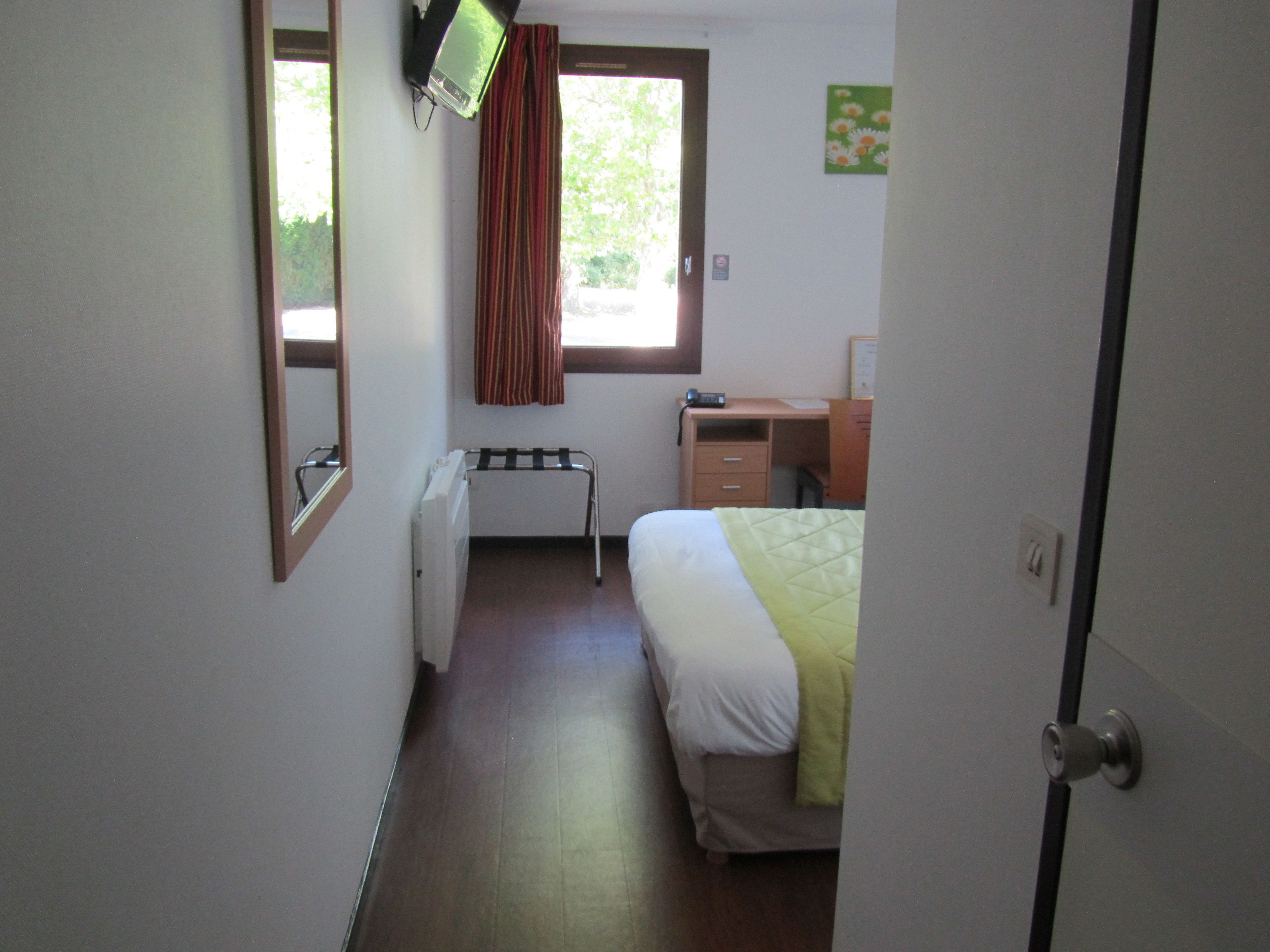 A bedroom with a bed, a desk, a chair, a radiator, a window with curtains, and a television mounted on the wall.
