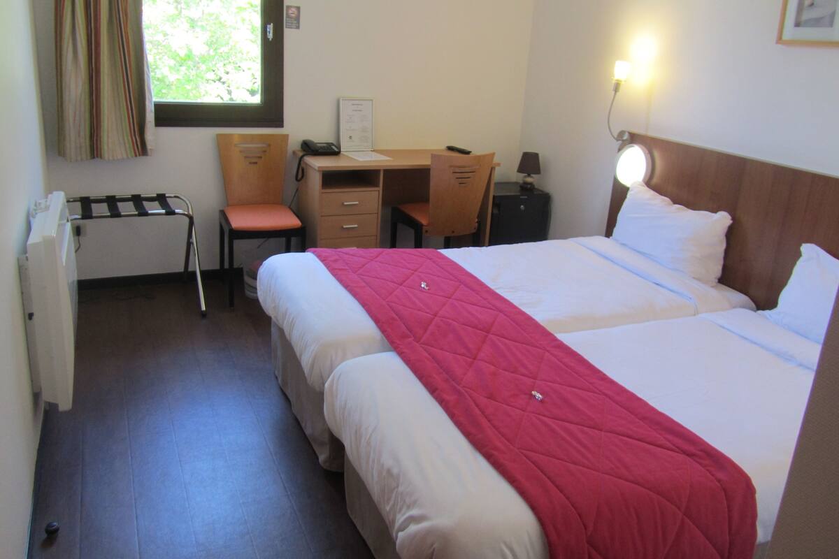 Habitación de hotel con cama, escritorio, silla, radiador y ventana con cortinas.
