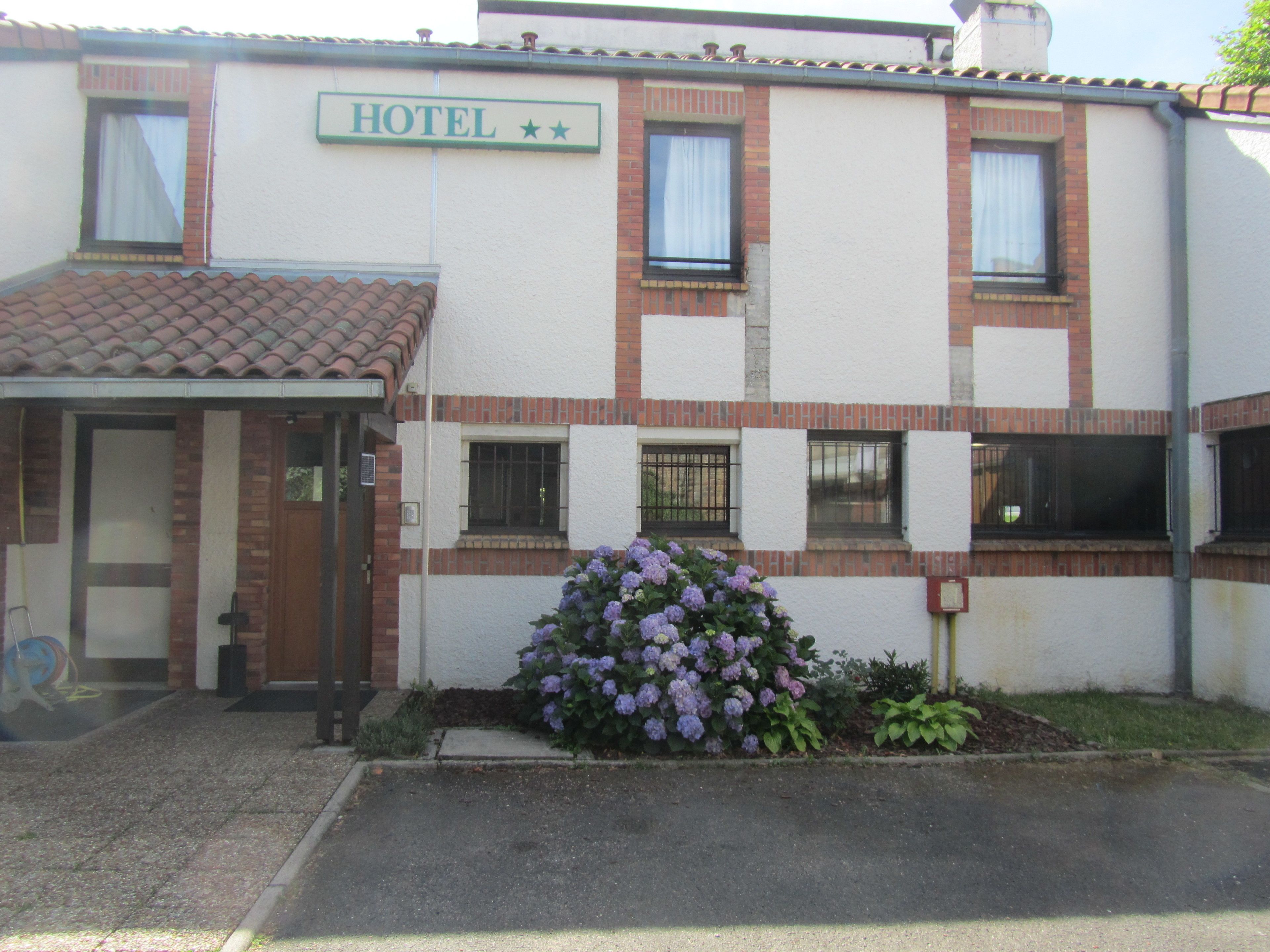 A hotel with a sign reading "HOTEL" above the entrance, a covered entrance, and a small garden with purple flowers.