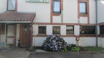 A hotel with a sign reading "HOTEL" above the entrance, a covered entrance, and a small garden with purple flowers.