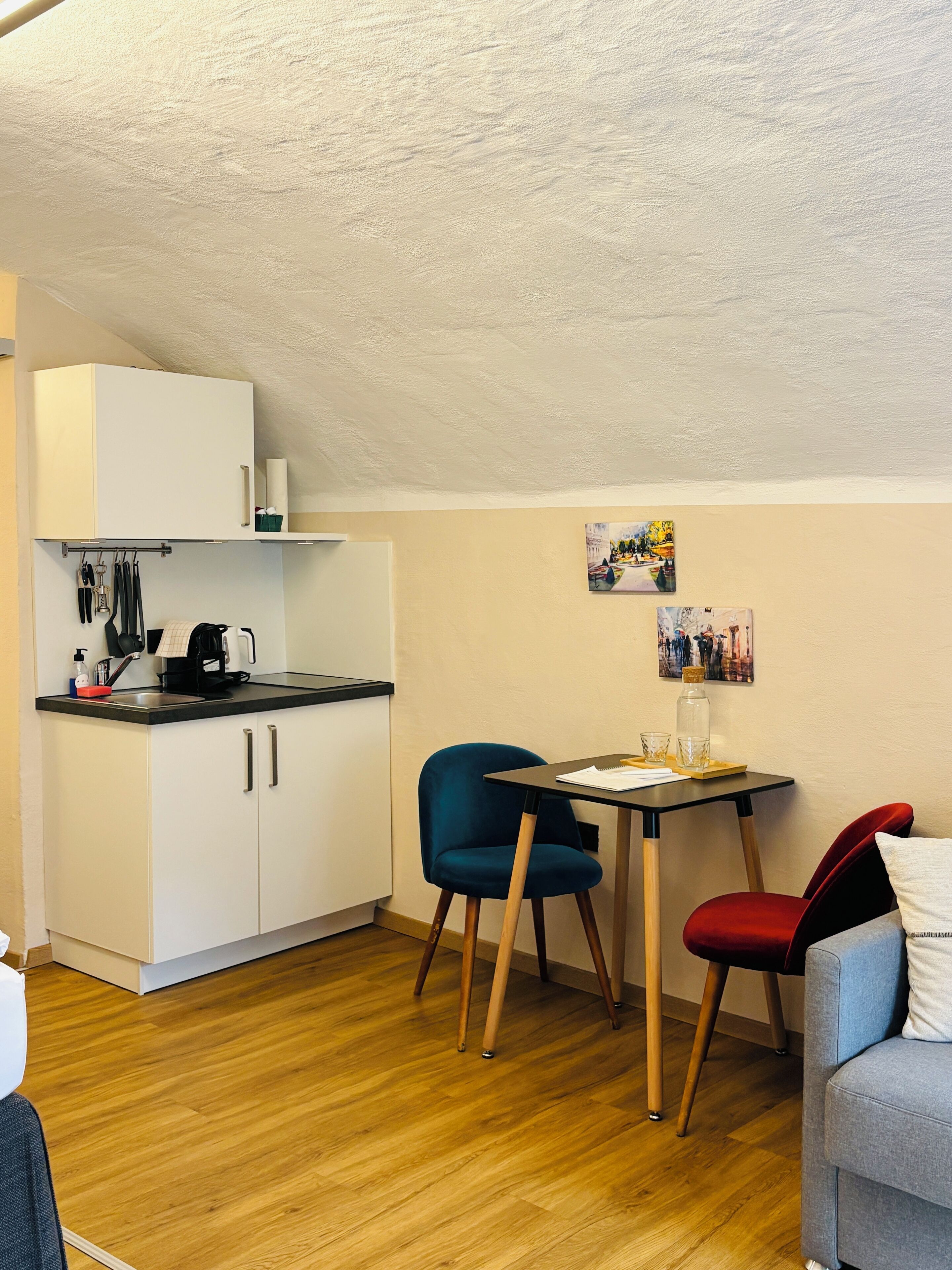 A compact kitchen with white cabinets, a black countertop, and a coffee machine. There is a small dining table with two chairs, one blue and one red.