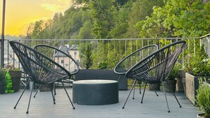 An outdoor seating area with two black chairs and a round table on a wooden deck, surrounded by potted plants and overlooking a lush green hillside.