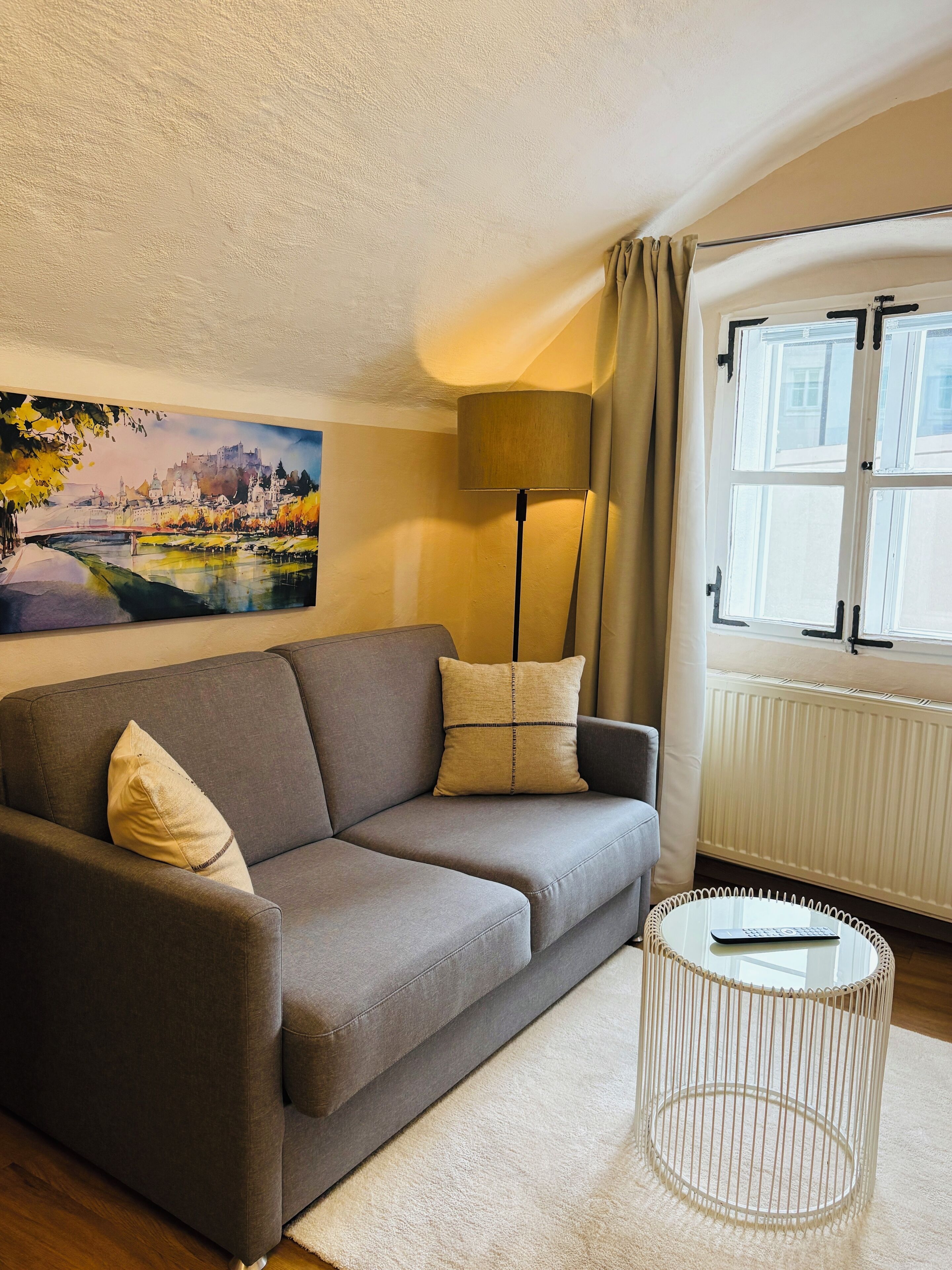 A living room with a grey sofa, beige cushions, a floor lamp, a painting of a cityscape, and a window with curtains.
