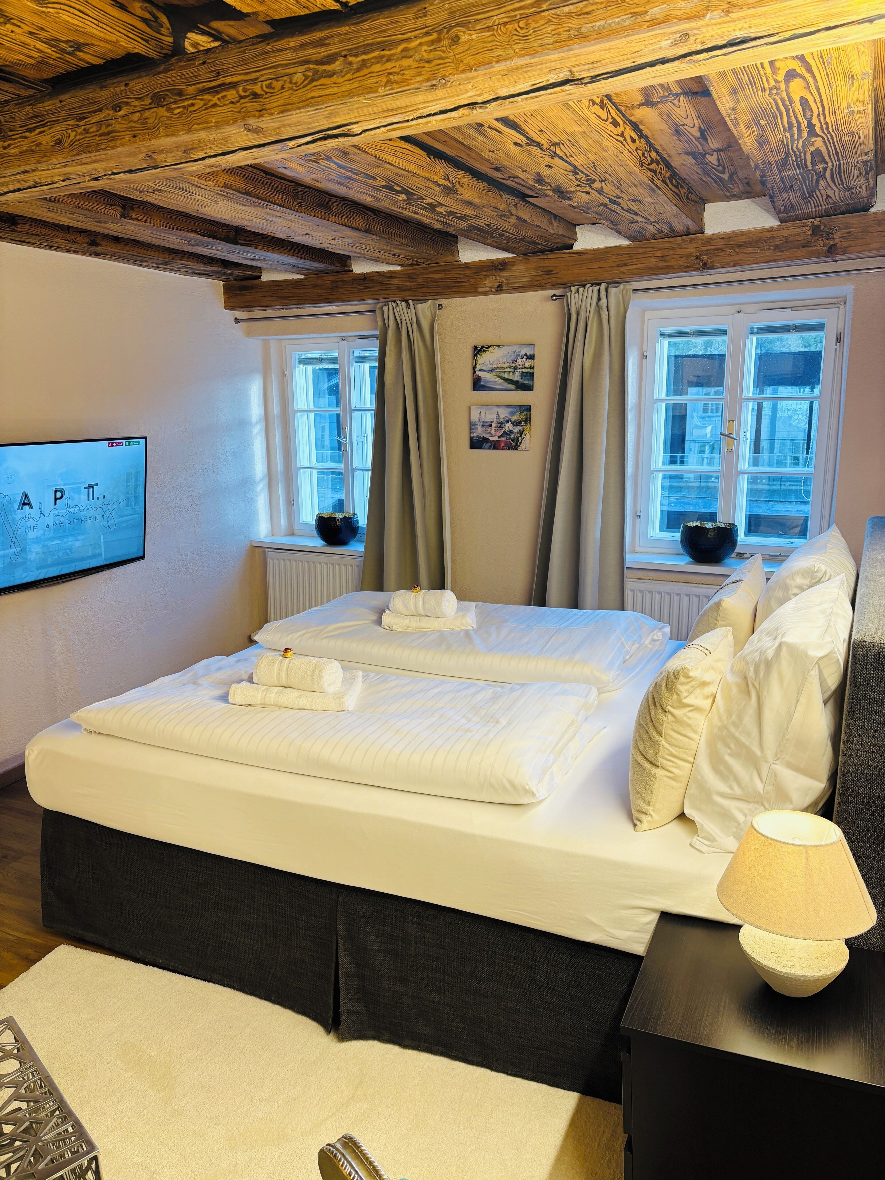 Un lit bien fait, avec du linge de lit et des serviettes blancs, une table de chevet noire avec une lampe, et un plafond en bois.