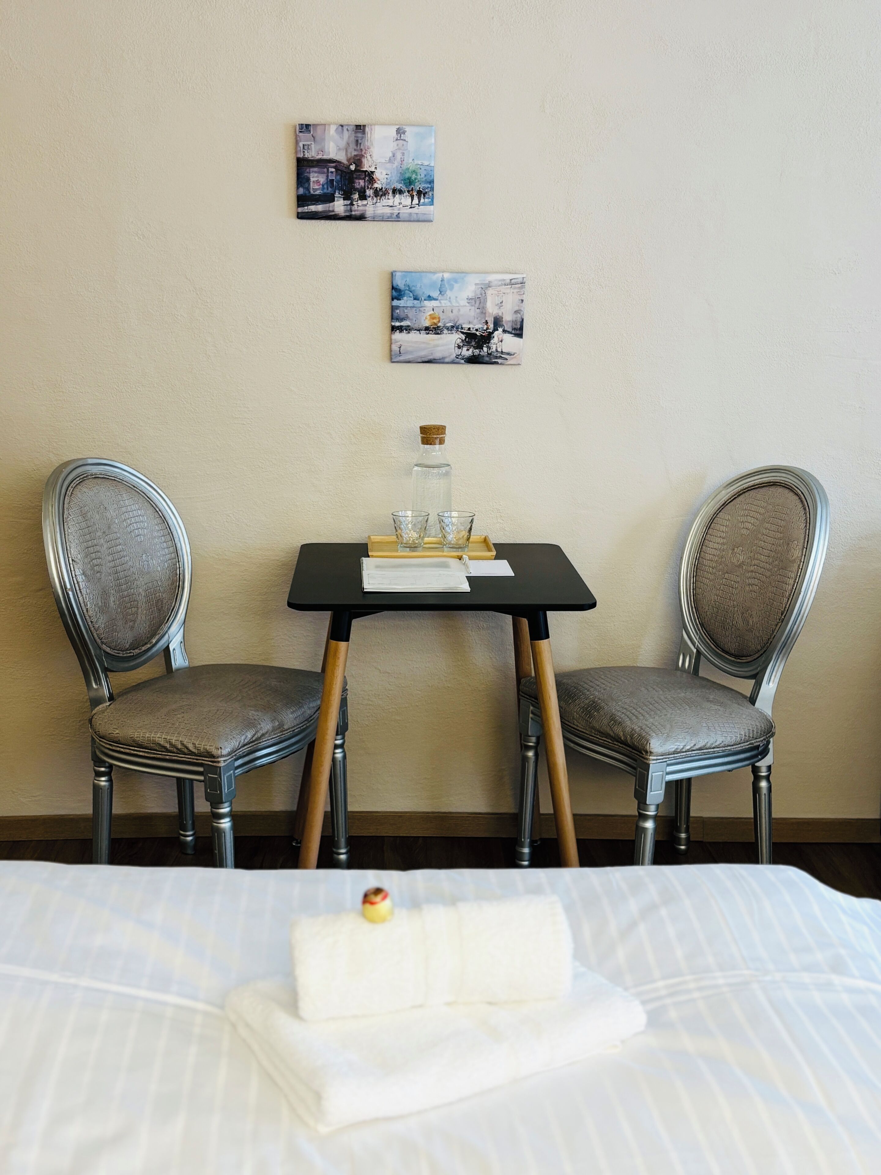 Et hotelværelse med to polstrede lænestole, et lille bord med en flaske og glas samt en seng med et pænt foldet hvidt håndklæde.