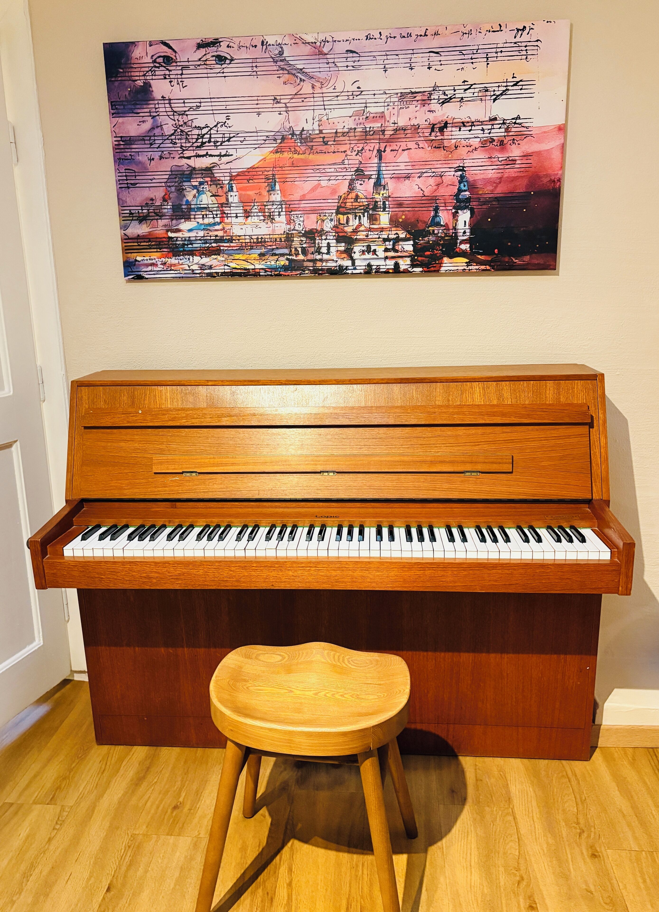 A piano with a stool in front of it, a wall with a colorful painting above.